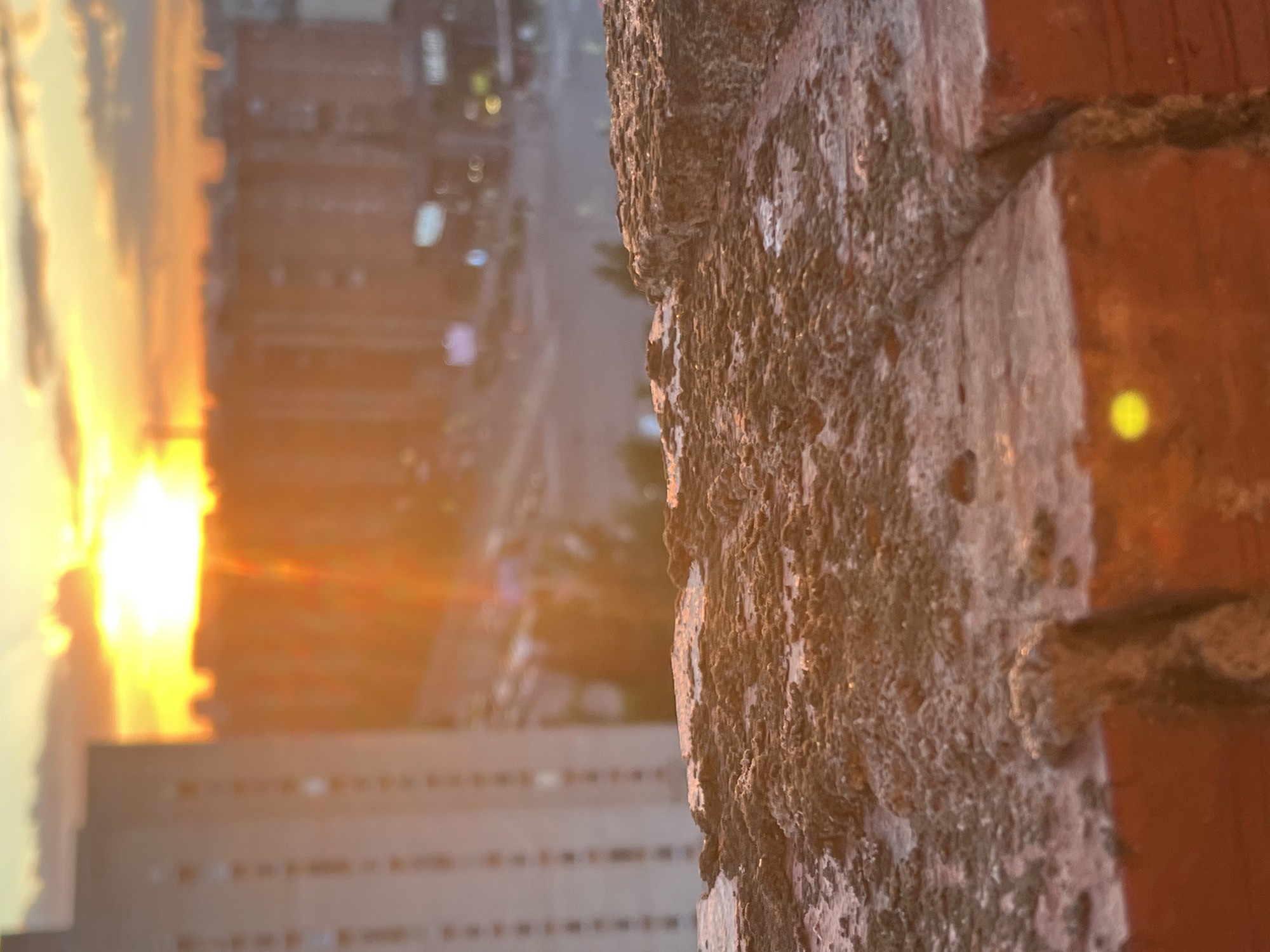 The media file shows a close-up of a rough, textured brick surface in the foreground, which occupies the right side of the frame. The brick appears weathered, with visible mortar and an uneven, pitted texture. A glint of light, possibly a reflection or a light source, is visible on the brick.
In the background, a city scene is visible, out of focus and bathed in warm, golden light characteristic of either sunrise or sunset. The light creates a strong lens flare effect, particularly on the left side of the image, where the sun appears to be setting or rising behind a vertical structure. Silhouetted against this bright light is a tall, rectangular building, likely an apartment complex, with multiple rows of windows. Beyond this building, more city structures and lights are discernible, suggesting an urban environment. The overall impression is one of a dramatic sunset or sunrise over a city, viewed from a slightly elevated position, with the foreground focus on the textured brick. There is no visible text in the image.