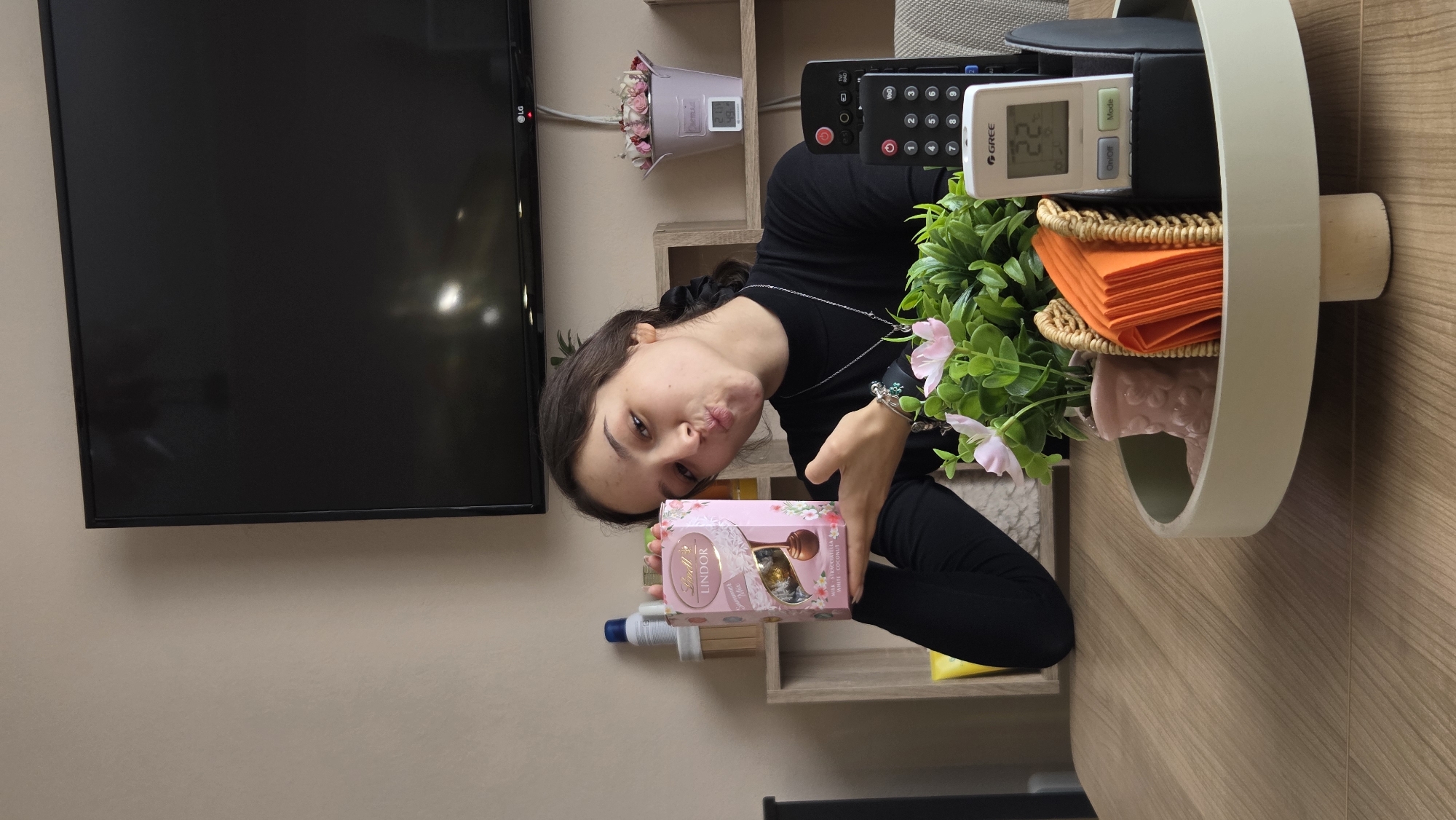 The image depicts a young woman indoors, holding a box of Lindor chocolate candies with a puckered expression. She is wearing a black top and stands adjacent to a shelf unit mounted on a light beige wall. The shelf unit houses a small bouquet of flowers in a pink pot, a black remote control, a Gree air conditioning remote, and other miscellaneous items. To the left of the shelf unit is a black LG flat-screen television mounted on the wall. A round, pale-green side table stands to the right of the woman, adorned with faux foliage and orange napkins. The background is a wall and part of a floor with a wood-grain pattern. There is no information to confirm or deny the location is in the city of Enina, Bulgaria.