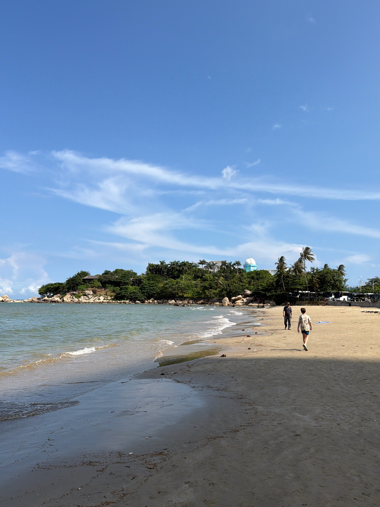 The image depicts a beach scene in Nha Trang, Vietnam, under a bright blue sky with scattered white clouds. The foreground features a sandy beach with gentle waves lapping at the shore. The water is a calm, greenish-blue.
Further back, a rocky outcrop covered in lush green trees and vegetation forms a headland. On top of this headland, several buildings and a distinctive blue dome structure are visible, suggesting a research facility or observatory. Palm trees are interspersed with the other foliage, characteristic of a tropical coastal environment.
Two individuals are walking on the beach. One person, wearing a dark shirt and pants, is closer to the water and walking away from the viewer. The second person, wearing a patterned shirt and shorts, is further up the beach and also walking away from the camera. The sand shows signs of recent wave activity, with damp patches and some debris. In the background, a line of white buses parked along a road suggests a nearby parking area or transport hub. The overall atmosphere is one of a serene and sunny day at the beach.