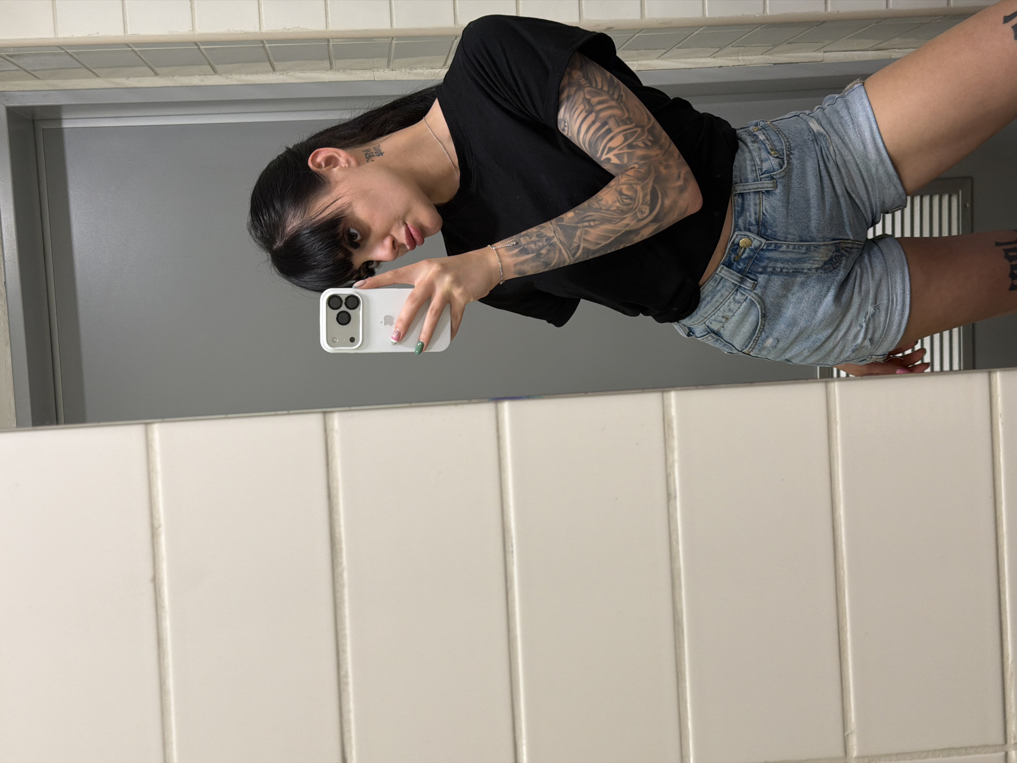 An indoor mirror selfie captures a young woman standing in what appears to be a bathroom or a tiled hallway in Plovdiv, Bulgaria. The woman has long, dark hair with bangs and striking full sleeve tattoos on both arms, featuring intricate designs. A small tattoo with partially visible text, "Don't worry about," is seen behind her left ear. She wears a plain black short-sleeved t-shirt and light blue distressed denim shorts. On her right thigh, a tattoo partially reads "real."
She is holding a white iPhone with a triple-lens camera setup, using it to take the photo. Her left hand shows green nail polish, while her right hand has glittery pink nail polish, and she wears a delicate silver bracelet. A thin silver chain necklace is also visible around her neck. She looks directly at the phone's camera with a subtle smirk.
The background, reflected in the mirror, shows a simple setting. The lower portion consists of white rectangular ceramic tiles with light-colored grout lines, suggesting a wall or wainscoting. Above the tiles, a plain grey surface, possibly a wall or door, is visible. The lighting is bright and even, characteristic of an indoor environment.