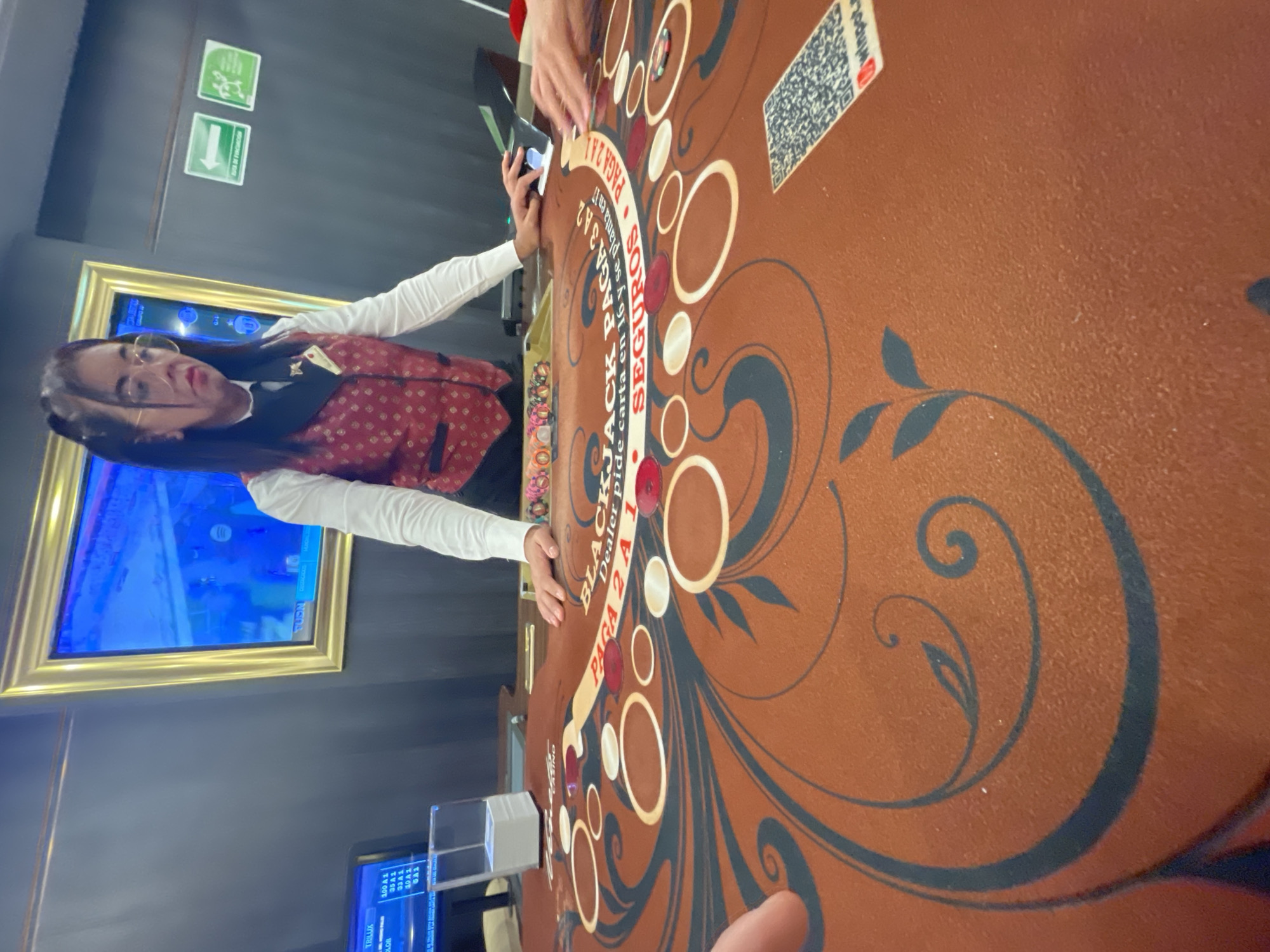 The image shows a woman in a red vest with a white long-sleeved shirt dealing cards at a blackjack table. The table is brown with a circular design and text that reads "BLACKJACK", "PAGA 2 A 1", and "SEGUROS". There are chips on the table. In the background, there is a TV screen on the wall, and a smaller screen is visible below the table. Signage on the wall indicates emergency exits. The text "RUTA DE FIRCOMCIGE" is visible, as well as information about a brand called "TRILUX" and "winpot". The location can be identified as being in Huixquilucan de Degollado, Mexico, based on the Spanish text on the table.