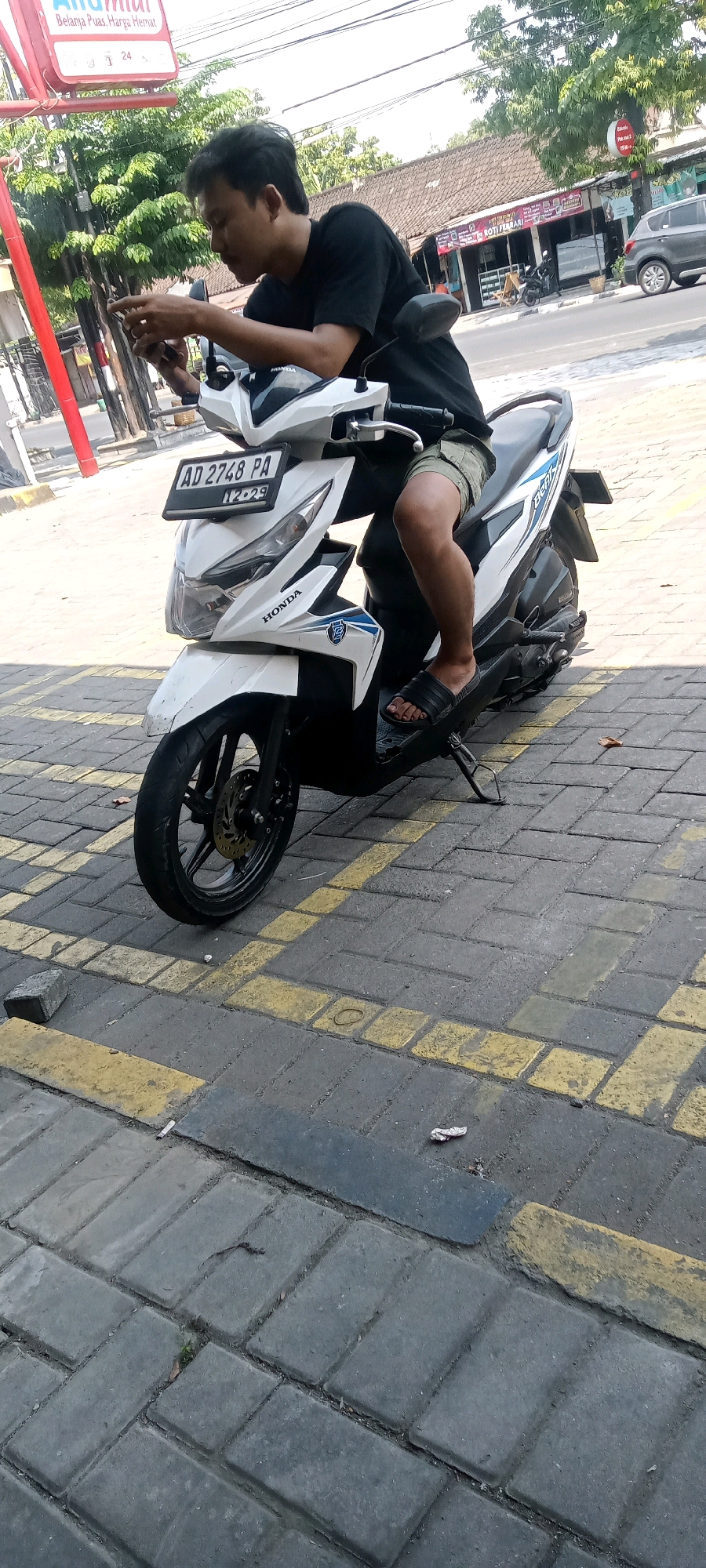 The image shows a young man sitting on a white Honda Beat scooter parked on a cobblestone sidewalk with yellow painted parking lines, likely in the city of Surakarta, Indonesia. The man is wearing a black t-shirt and olive green shorts and is looking down at his smartphone. The scooter has a license plate reading "AD 2748 PA". In the background, there are shops and trees. The scene appears to be on a bright, sunny day.