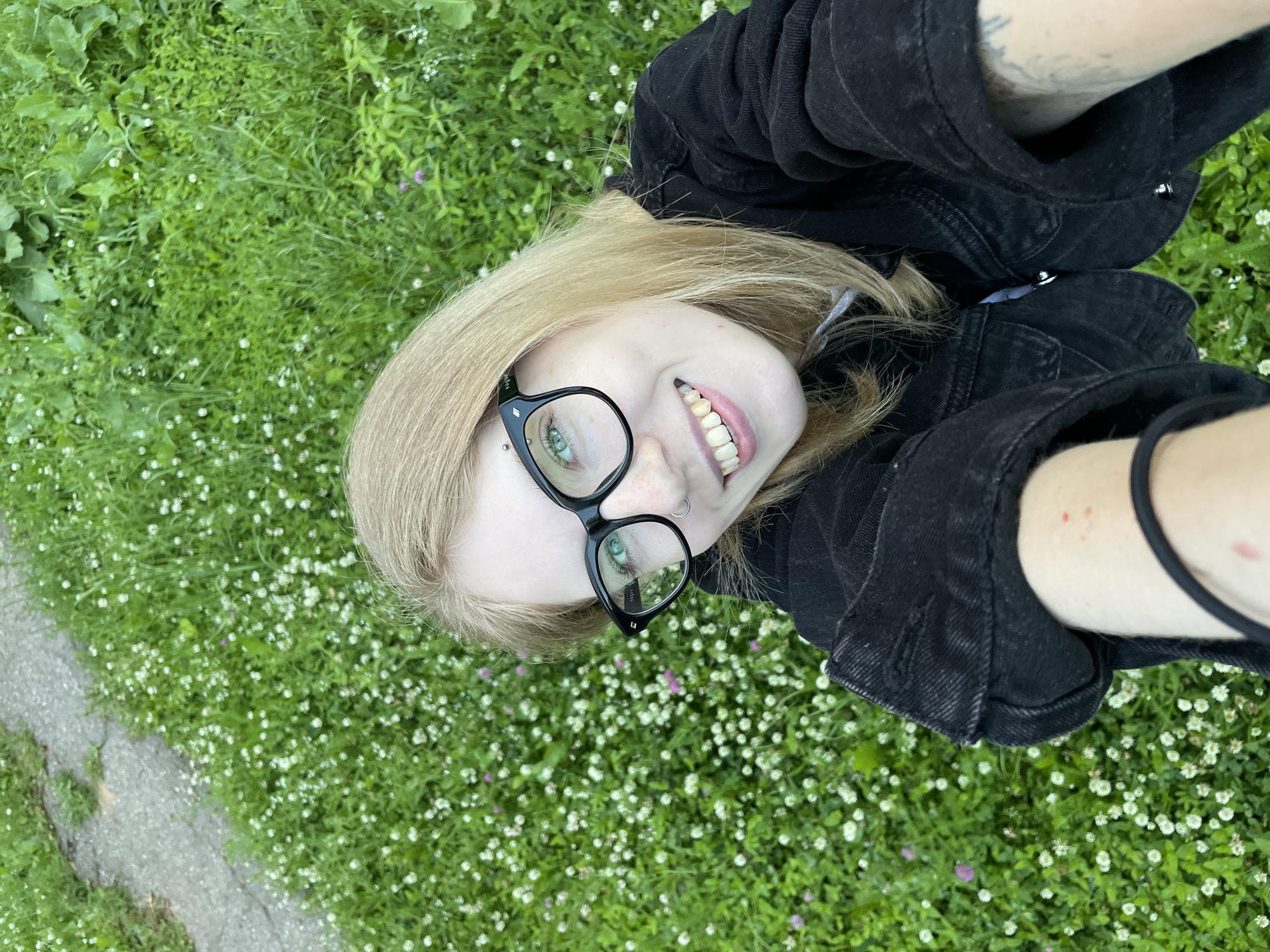 The image is a close-up, high-angle selfie of a young person lying on a grassy area dotted with small white and purple wildflowers. The subject has fair skin, blonde hair, and is wearing black-rimmed glasses and a black denim jacket. They are looking directly at the camera with a smile, revealing teeth. A nose ring is visible. Tattoos can be seen on their left arm. The grassy area appears to be an outdoor setting, possibly a park or field, with a paved path visible to the left of the frame. The lighting suggests it is daytime, likely a clear or partly cloudy day, given the bright illumination. The overall mood seems casual and relaxed. No visible text is present in the image. The location context is Izhevsk, Russia.