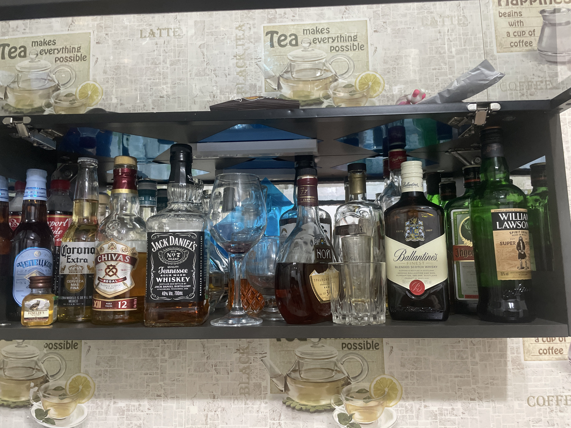 The image shows a shelf stocked with various alcoholic beverages, including vodka, whiskey (Jack Daniel's and William Lawson's), beer, and scotch (Chivas Regal and Ballantine's). The shelf also contains glasses. The backdrop is a wall with images related to tea and coffee. Text on some of the bottles is written in Russian. Some of the bottles are labeled as being produced in Ukraine and Armenia. The precise location is unknown, but is within Russia as indicated by the presence of Cyrillic lettering.