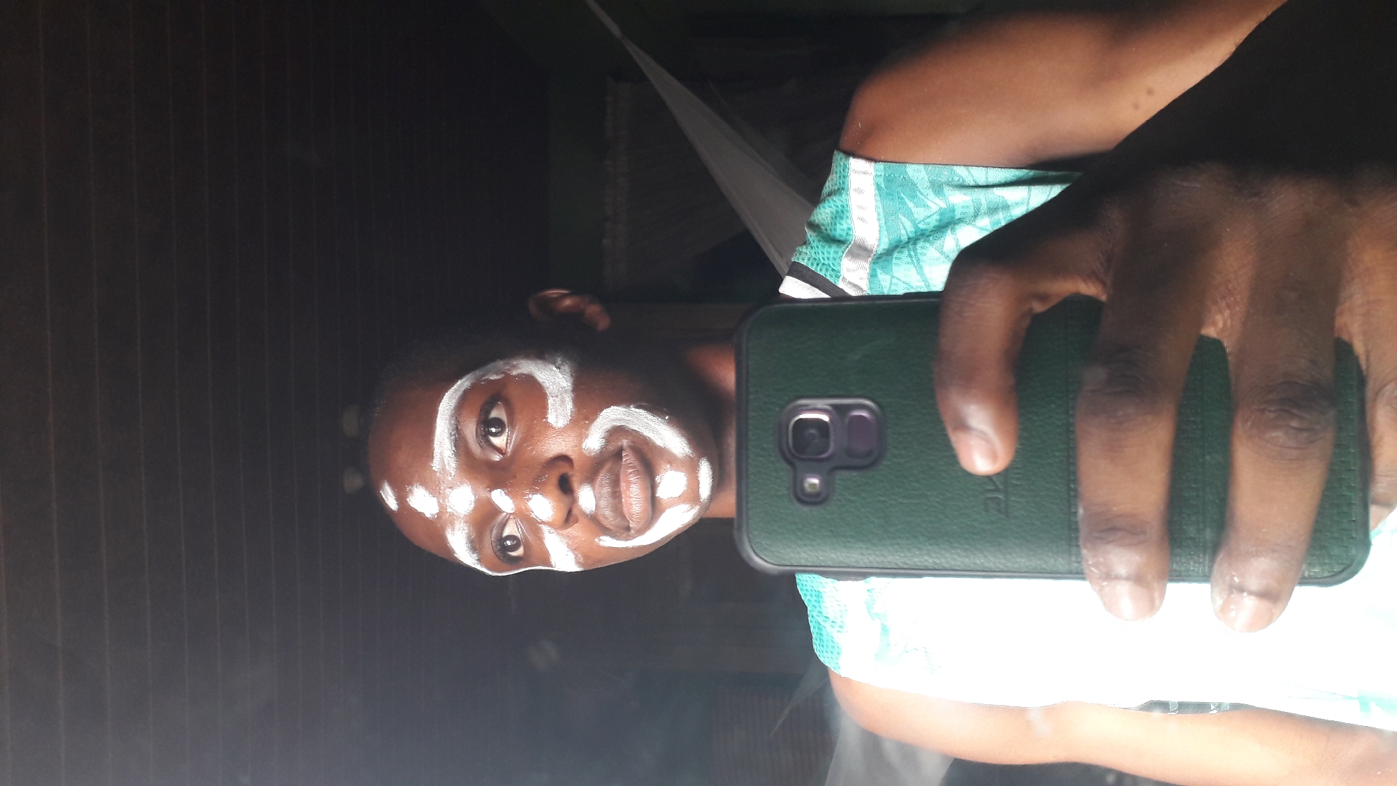 A person with white markings on their face, resembling tribal or ritualistic paint, takes a selfie with a green-cased mobile phone. The background includes a wooden-paneled wall and what appears to be a mosquito net. The location is inferred to be Nkpor, Nigeria.