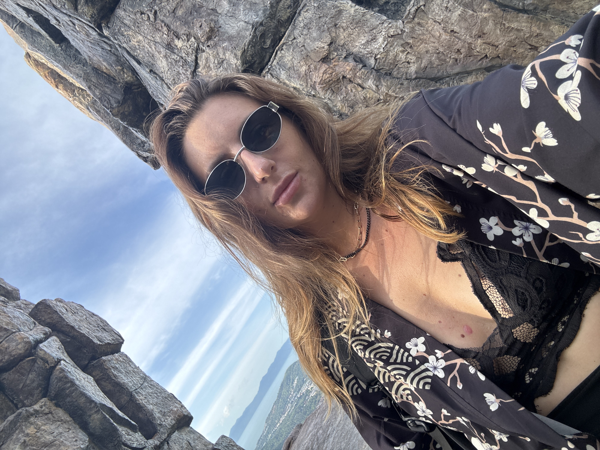 The media file is a portrait-oriented, selfie-style photograph of a woman. She is wearing sunglasses, a black kimono-style jacket with a white floral pattern, and a black lace bra. Her long, blonde hair is down and frames her face. She is wearing a necklace. The background consists of large, grey rocks and a blue sky with wispy white clouds. In the distance, a body of water and a city or town are visible, suggesting a scenic, elevated location. The lighting indicates it is daytime, possibly late afternoon given the warm tones. The woman's expression is neutral, and the overall mood is relaxed and confident. There is no visible text in the image.