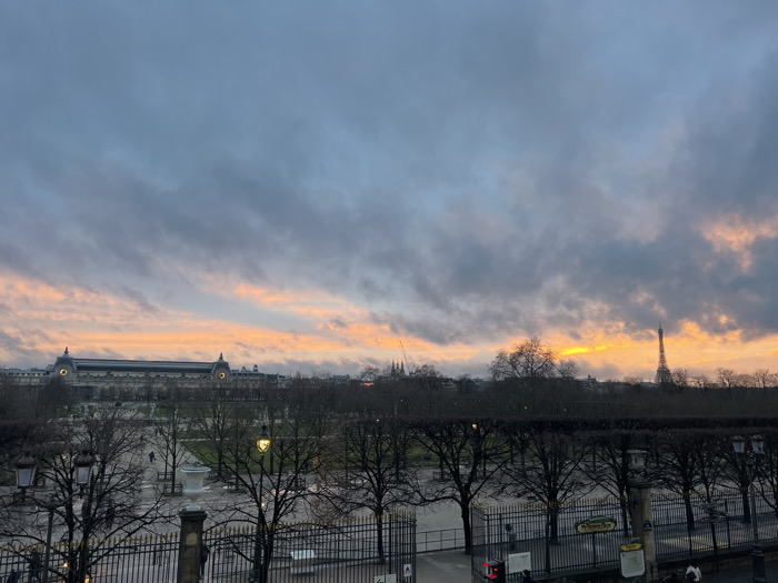 A panoramic view shows a park with wrought iron fences and bare trees under a cloudy sky. A strip of orange and pink light illuminates the horizon. Prominent structures include the Musée d'Orsay and the Eiffel Tower, barely visible in the distance through the trees. A "Métro" sign is visible near a park entrance. The scene suggests an early morning or late afternoon in Paris.