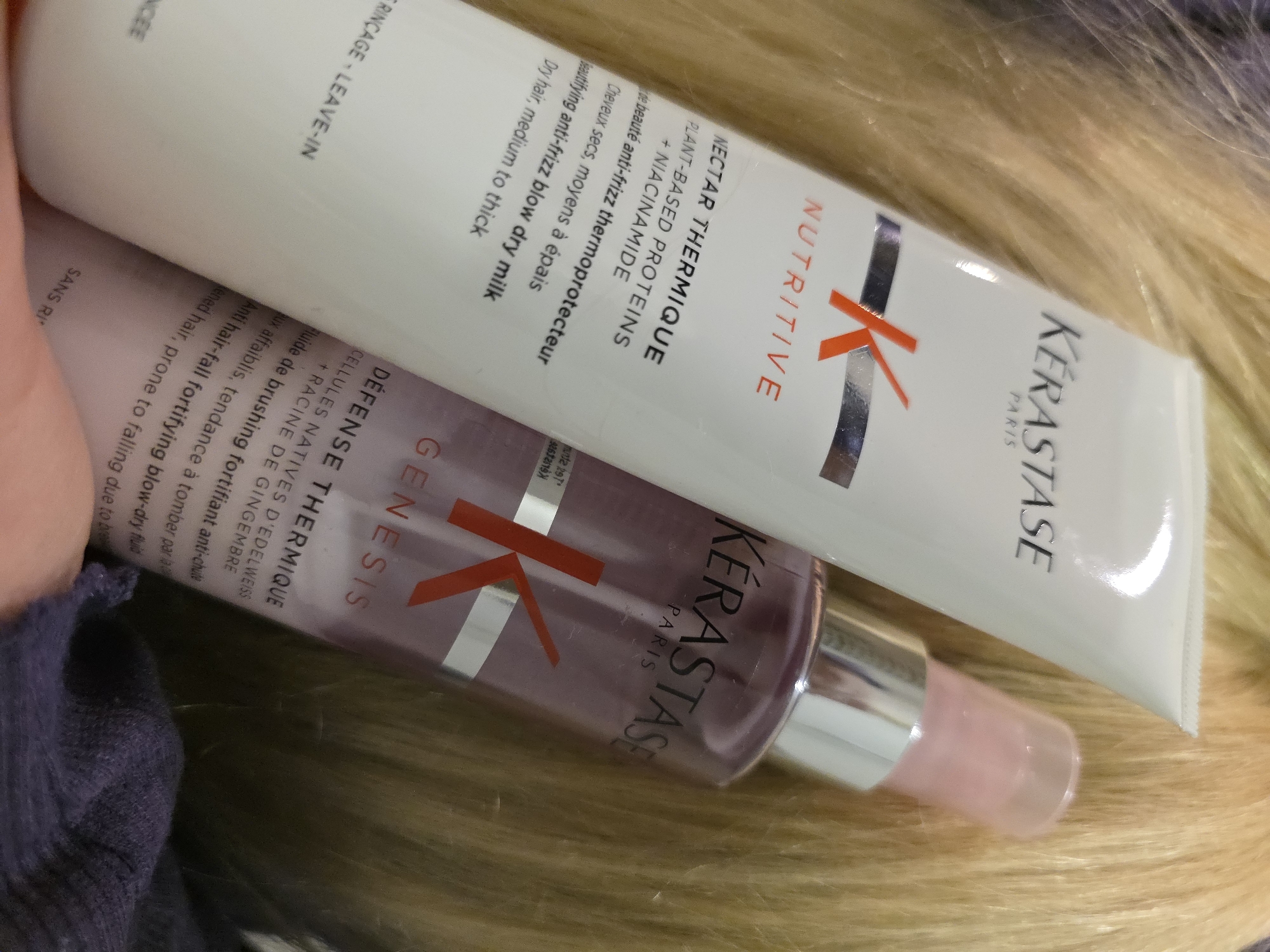 Here is a description of the image:
The image shows two Kerastase hair products placed on a background of blonde hair. One is the Kerastase Nutritive Nectar Thermique, described as a plant-based protein, anti-frizz blow dry milk for dry hair. The other is Kerastase Genesis Defense Thermique, an anti-hair fall fortifying blow-dry fluid with native edelweiss cells and ginger root, for weakened hair prone to falling due to breakage. The brand names are prominently displayed on each product.