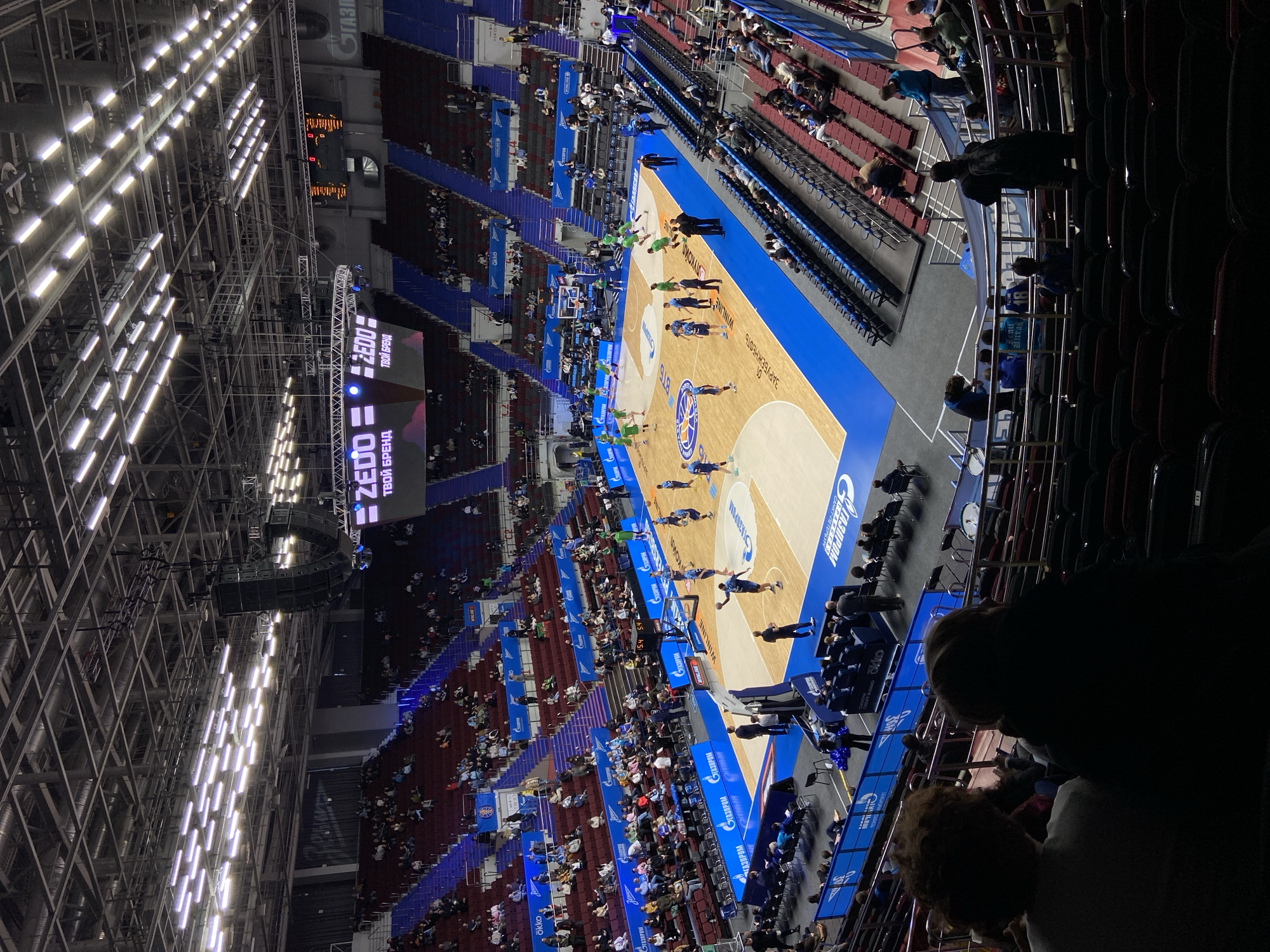 The image shows a basketball game at the Sibur Arena in Saint Petersburg, Russia. A game is in progress on the court, with players in blue and green uniforms. The arena is filled with spectators in the stands. The score is visible on the scoreboard with time remaining. A large ZEDO advertisement is seen above the court. Logos of Gazprom and other companies are displayed around the court. The arena's interior structure, including the lighting grid, is visible in the background.