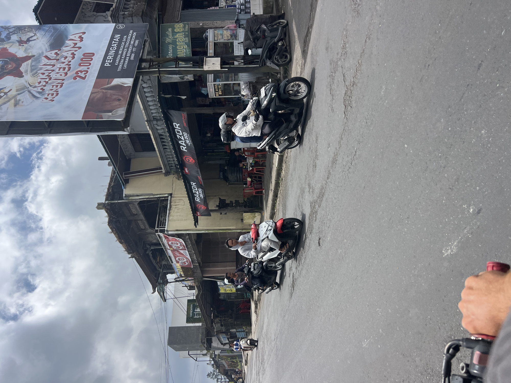 A street scene in Kintamani, Indonesia, shows several people riding motorbikes on an asphalt road. A large billboard with an advertisement and warning text is visible on the right side of the image. Several shops with awnings line the street, displaying banners and signs. Some of the shops appear to be food vendors, with tables and chairs set up outside. The sky is cloudy, and the overall atmosphere suggests a typical day in a busy town.