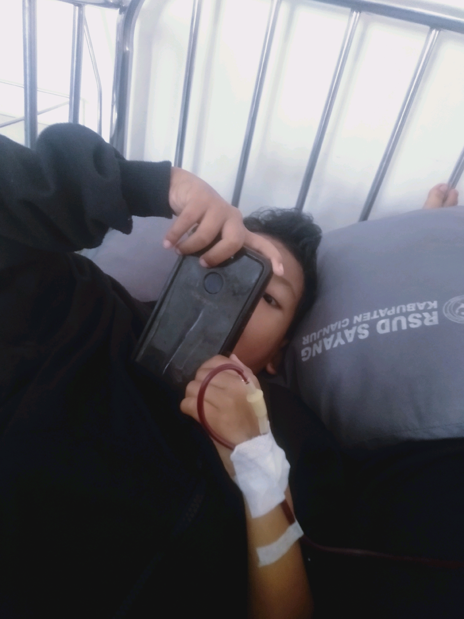 A young person is lying down, possibly in a hospital bed, holding a smartphone and looking towards the camera. They have a blood transfusion line attached to their arm, which is secured with white medical tape and gauze. The person is wearing a dark long-sleeved shirt. Behind them, a gray pillow with the text "RSUD SAYANG KABUPATEN CIANJUR" is visible. The background shows the metallic bars of a bed frame and a white wall.