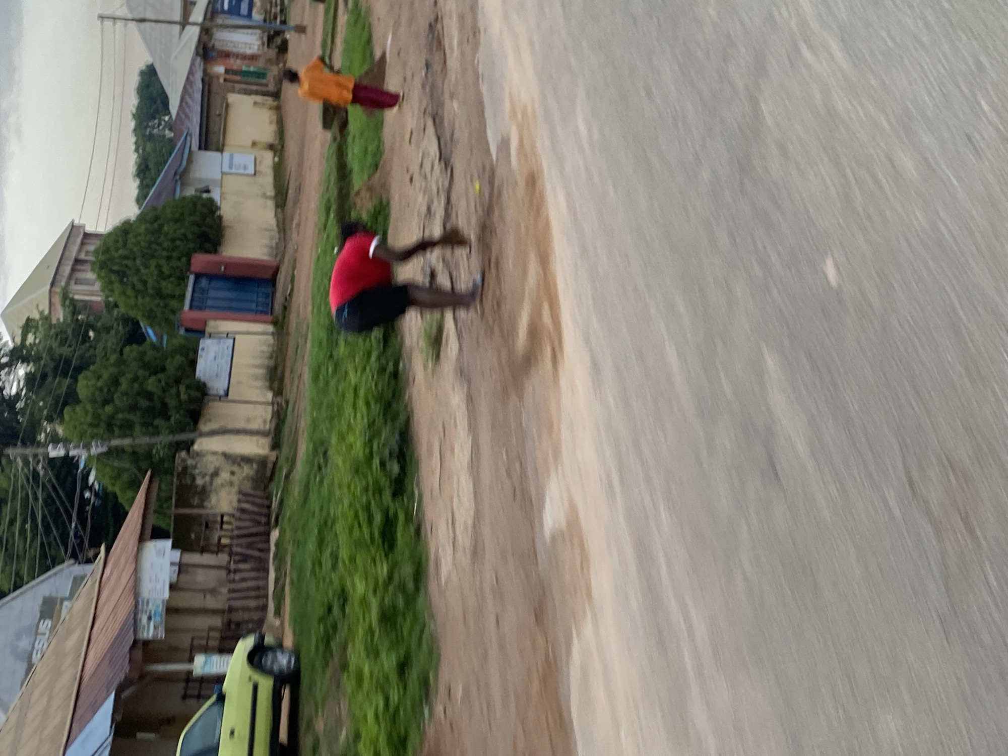 The image depicts a street scene in Jos, Nigeria. In the foreground, a person in a red shirt and black shorts is bent over, appearing to sweep or clear the dirt road. Further down the road, another person in an orange shirt and maroon pants is walking away from the camera. The street is unpaved and appears rough, with patches of greenery along the side. Buildings are visible in the background, some with corrugated metal roofs. A yellow car is parked on the left side of the road near one of the buildings. The sky is overcast.