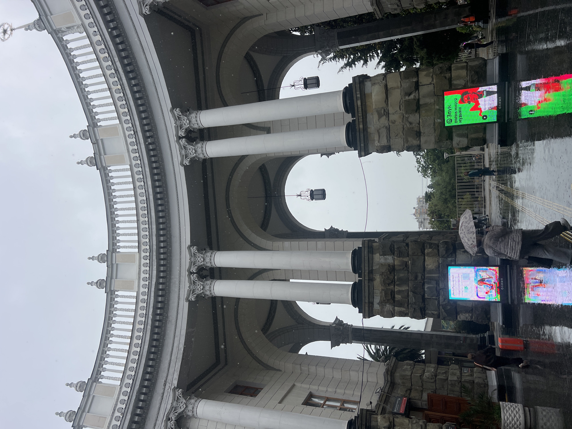 The image depicts a street scene in Sochi, Russia, taken on a rainy day. The architecture features a grand, arched structure with white columns, ornate details, and arched openings. The columns are set on stone bases. The sky is overcast, and rain is visible. Modern LED screens are mounted beside the columns. Pedestrians with umbrellas are seen in the foreground and background of the image. A person with a red suitcase is on the right-hand side of the frame. The ground is wet, reflecting the city lights.
