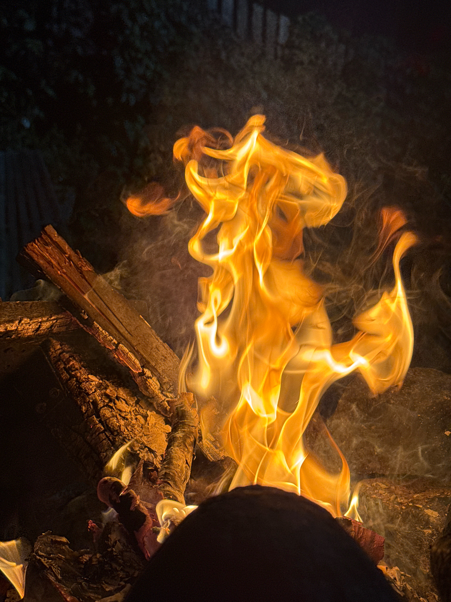 The image shows a close-up of a bonfire with flames in varying shades of orange and yellow, interspersed with wisps of smoke against a dark, out-of-focus background. Several pieces of wood are visible, some charred and broken, forming the base of the fire. The scene appears to be outdoors, possibly in a backyard or park in Kempelevo, Russia, given the context. The lighting is low, with the fire being the primary light source, casting a warm glow on the surrounding elements. No people are clearly visible, though a dark, rounded object in the foreground may suggest a person's silhouette or an object placed in front of the fire. The overall atmosphere is one of warmth and natural ambiance, typical of a campfire setting.
