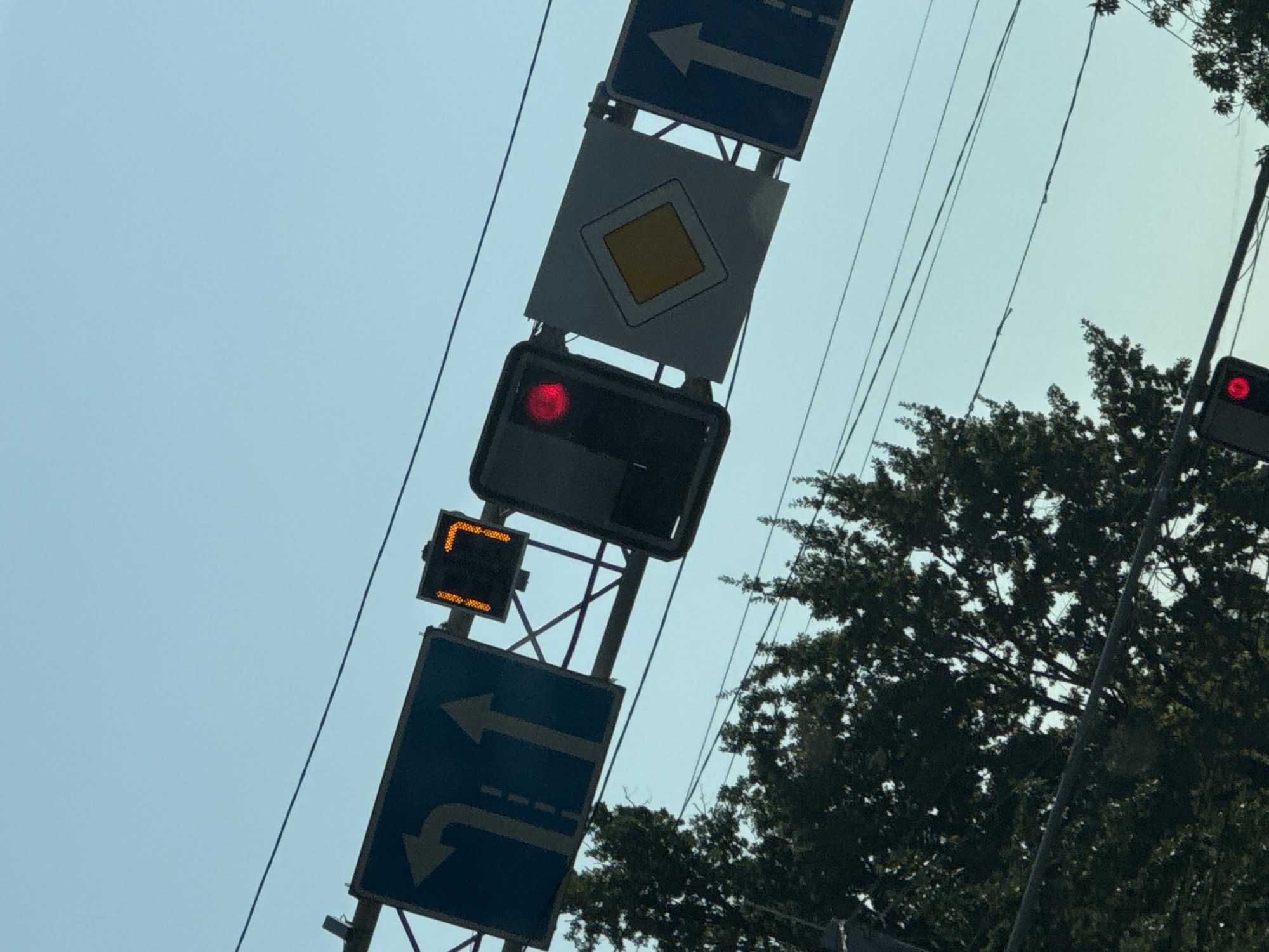 An overhead view shows a traffic light displaying a red signal, indicating a stop. Adjacent to the traffic light is a digital countdown timer showing "17" seconds. Below these are traffic signs. The leftmost sign indicates lanes for turning left and going straight. The sign to the right of the countdown timer is a diamond-shaped sign with a yellow interior, signifying a main road. Further to the right, another sign shows an arrow pointing straight ahead. The scene is set against a clear, pale blue sky. In the foreground, the tops of dark green trees are visible, suggesting a possibly urban or suburban environment. The image quality is fair, with some slight blurriness. The presence of traffic signals and signs points to an intersection in the city of Almaty, Kazakhstan.
