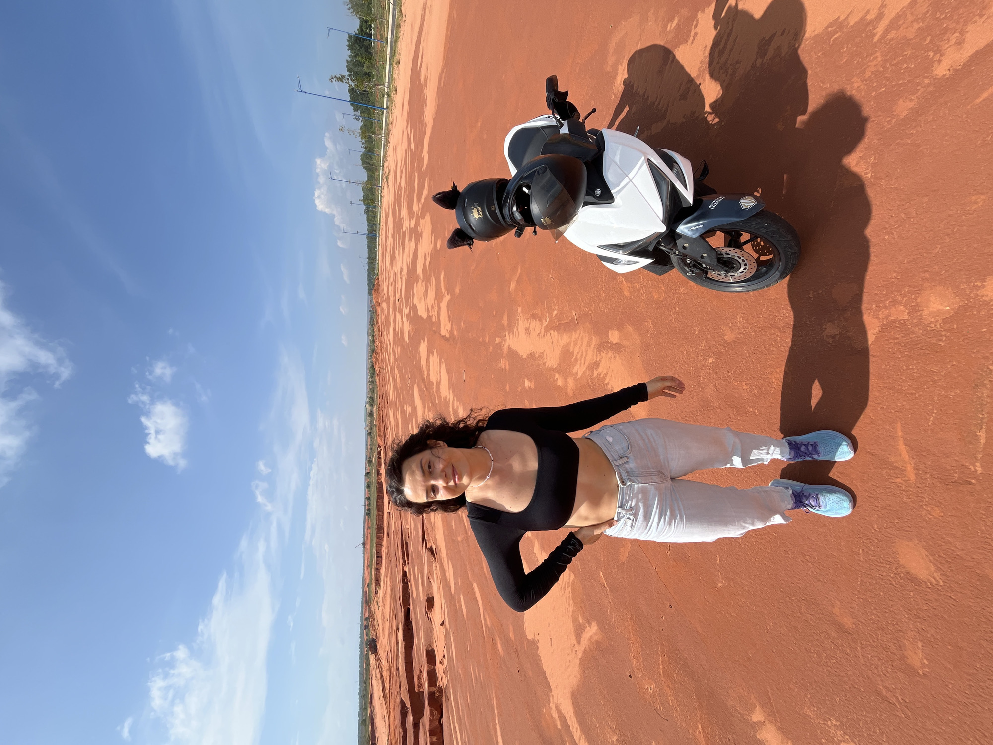A young woman stands in a red dirt quarry in Mui Ne, Vietnam, with a white scooter parked beside her. The woman is wearing a black long-sleeved crop top, light wash jeans, and blue sneakers. She has curly brown hair and is looking at the camera with a slight smile, her left hand on her hip. The scooter is white with black accents, and has two helmets on it, one of which has fluffy black cat ears. The ground is a vast expanse of red dirt with some uneven terrain and small eroded formations. In the background, there are some distant trees, power lines, and a clear blue sky with scattered white clouds. The scene suggests a sunny day in an outdoor, potentially arid, landscape.