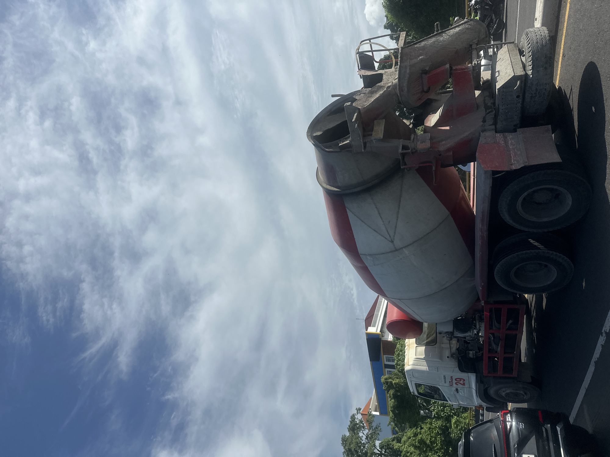 The image depicts a white and red cement mixer truck parked on the right side of a paved road in Denpasar, Indonesia, under a cloudy blue sky. The truck has the number "29" in red on its side. In the foreground, the rear of a black car is visible on the left. In the background, there are buildings with red roofs and trees. The scene suggests an urban environment, possibly a construction area or a street with moderate traffic. The lighting indicates it is daytime.