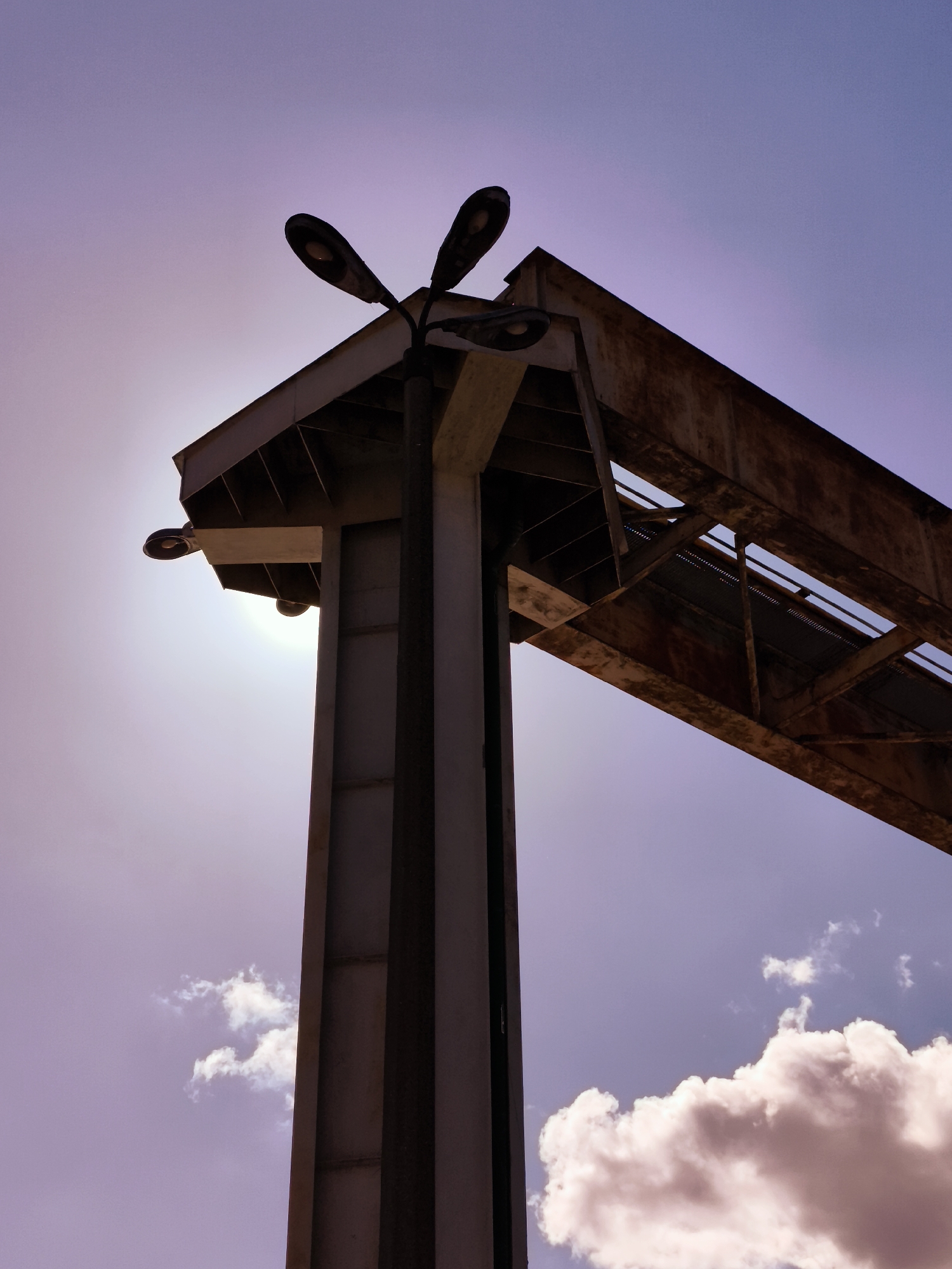 An upward, low-angle shot captures a portion of an industrial structure against a pale purple sky with scattered white clouds. A tall, dark, rectangular concrete pillar dominates the left side of the frame, supporting a cantilevered section of rusted metal superstructure. Three dark, modern streetlights are mounted on the pillar and the structure, with two positioned at the top, pointing outwards and upwards, and one more centrally located, pointing outwards. The sun is partially obscured by the pillar, creating a lens flare effect and silhouetting the overhead structure. The rusted metal appears to be part of a bridge or walkway, with visible beams and cross-bracing. The overall impression is one of an aged industrial landscape under a dusky sky, located in Gdańsk, Poland.