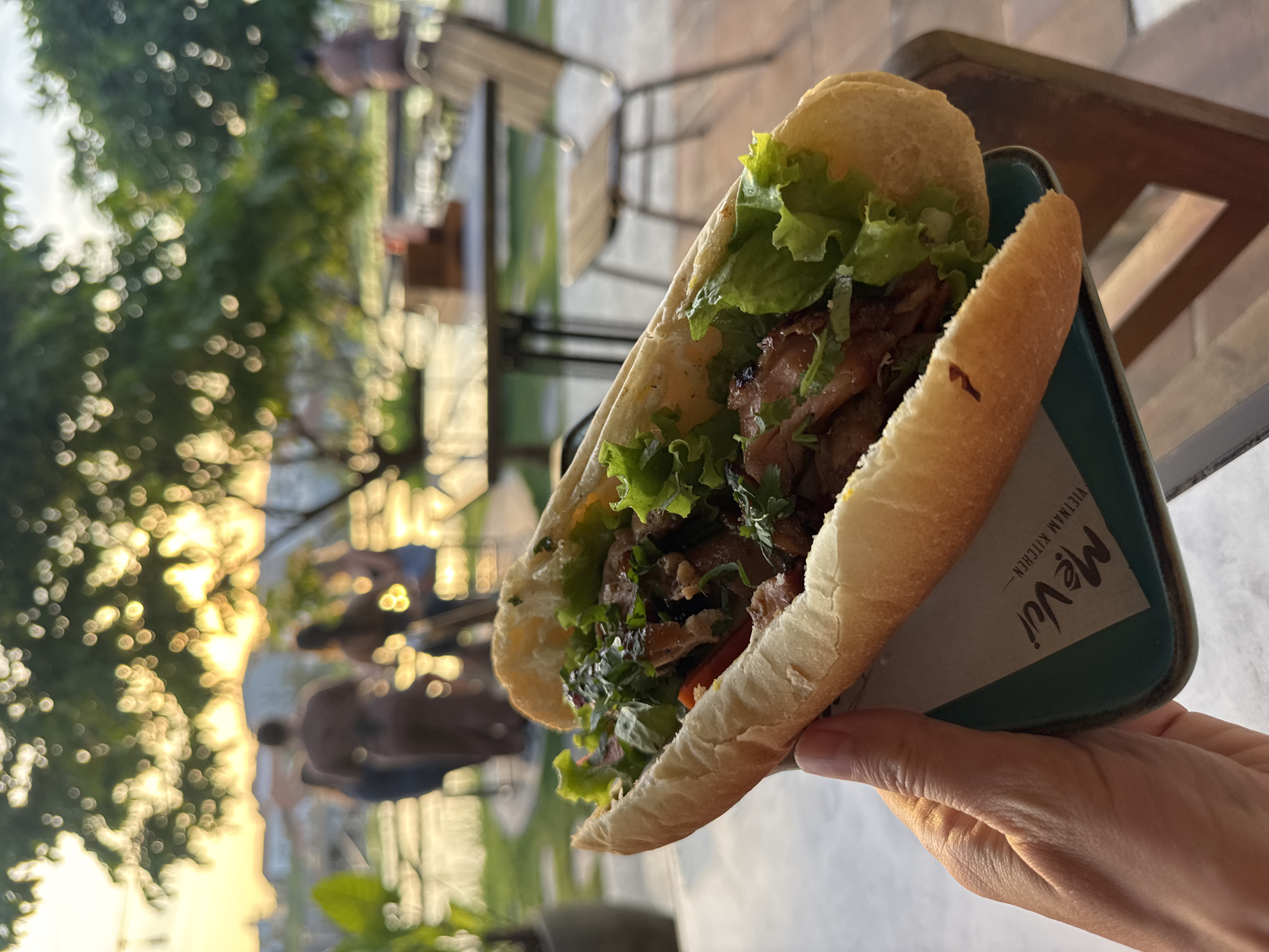 A close-up shot shows a person holding a Banh Mi sandwich in a serving container. The sandwich, filled with meat, lettuce, and herbs, sits in a dark teal container, which features the text "Mê Vui, Vietnam Kitchen." The background is blurred, but includes people sitting at outdoor tables, trees, and the setting sun. The location is North Kuta, Indonesia.