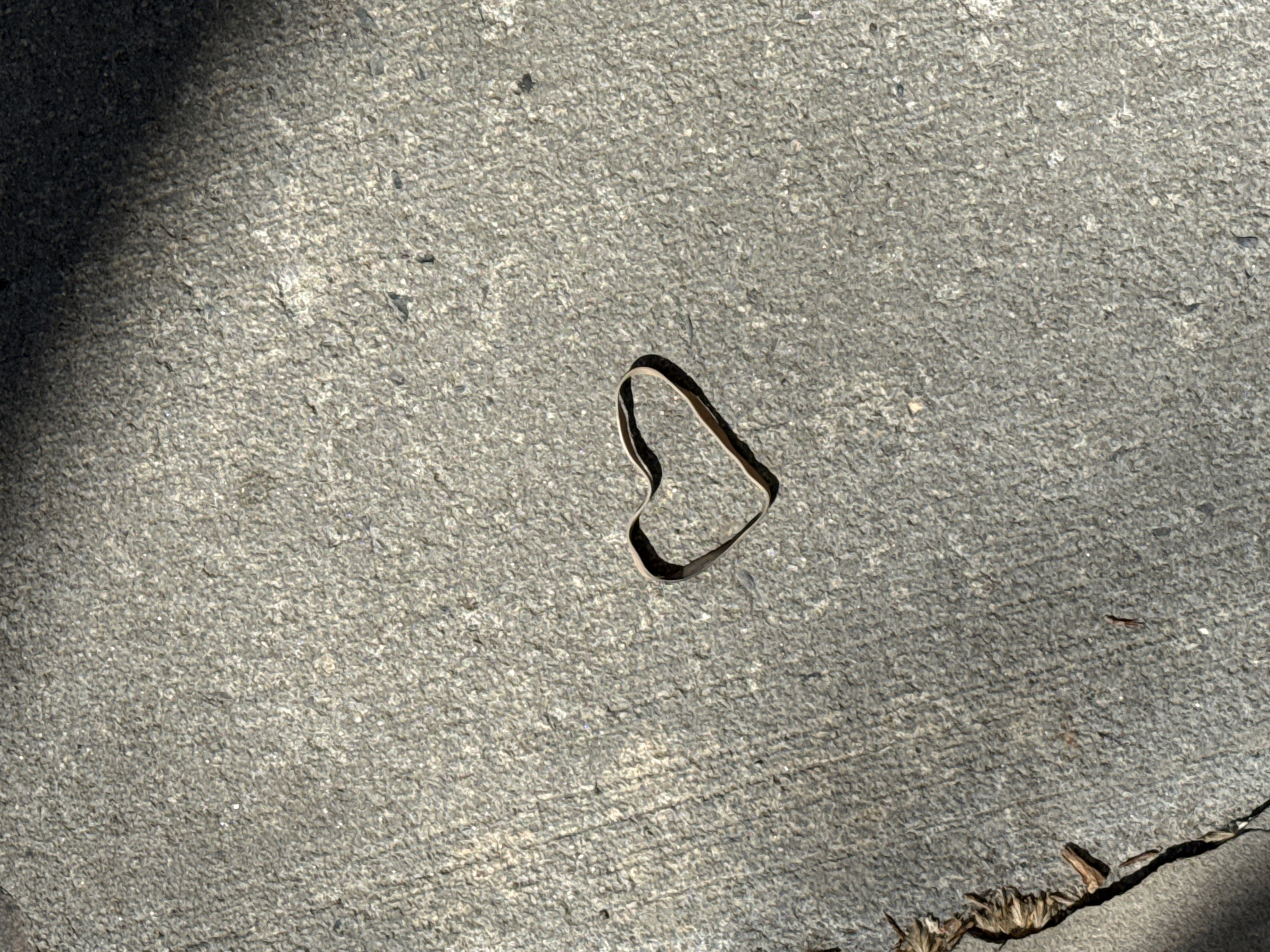 The scene is an outdoor concrete surface with a shadow in the upper left corner. In the center of the frame is a heart shape formed by a thin, dark object, possibly a piece of wire or material. The lower right corner has some debris, possibly dry leaves or twigs, along a crack in the concrete. The location is likely a sidewalk or paved area, but without further information, determining its precise location in New York City is not possible. There are no people visible.