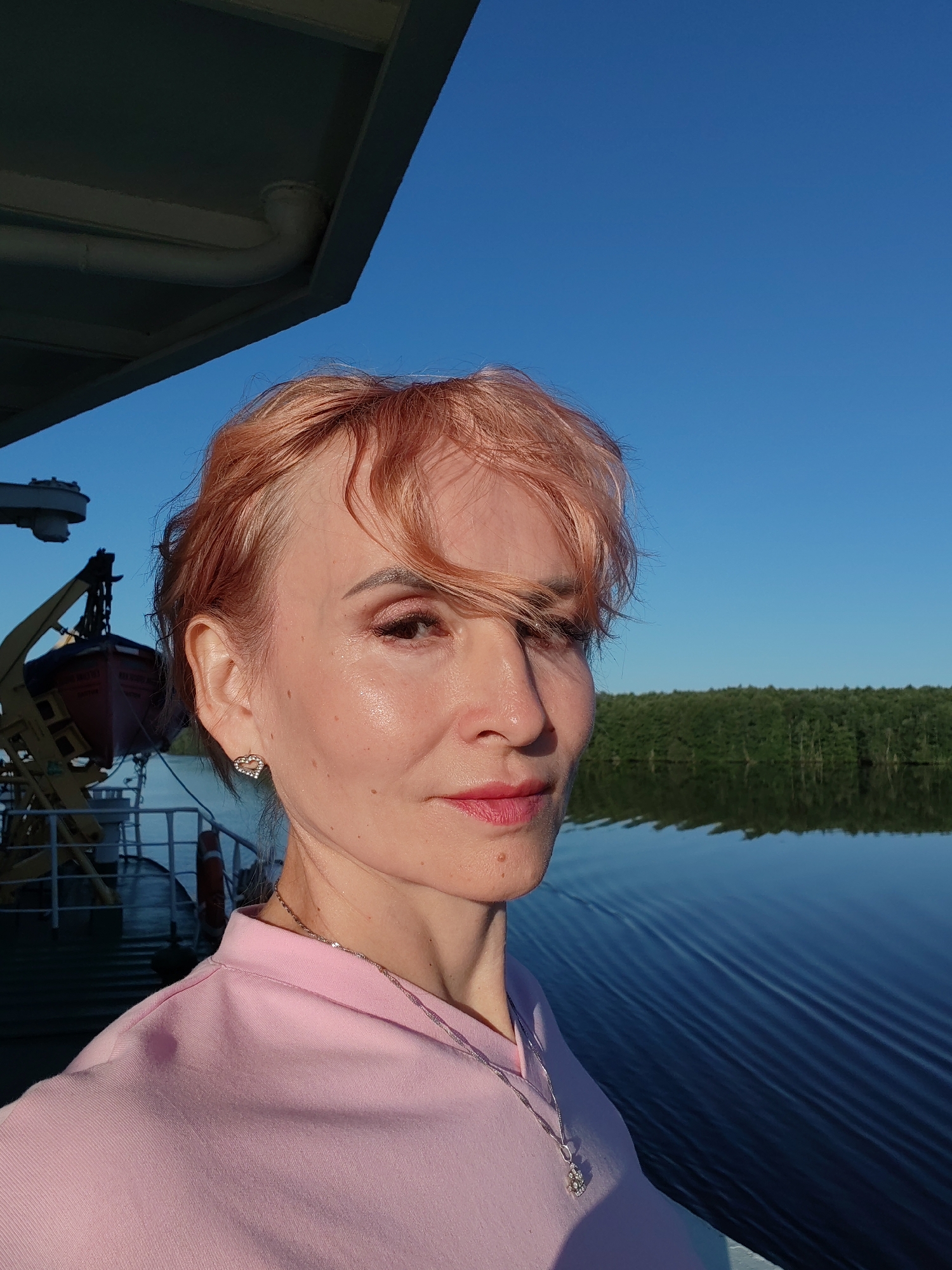 A woman with short, pink-hued hair and light makeup is taking a selfie on a body of water. She is wearing a pink shirt and a silver necklace with a pendant. She has a heart-shaped earring in her left ear. In the background, there is a green, tree-lined shore, a blue sky, and a body of water with ripples. To the left, part of a ship or boat structure is visible, including a life raft. The image appears to have been taken during daylight, with the sun casting a shadow on the woman's left side. The location is identified as being in Kurdyug, Russia.