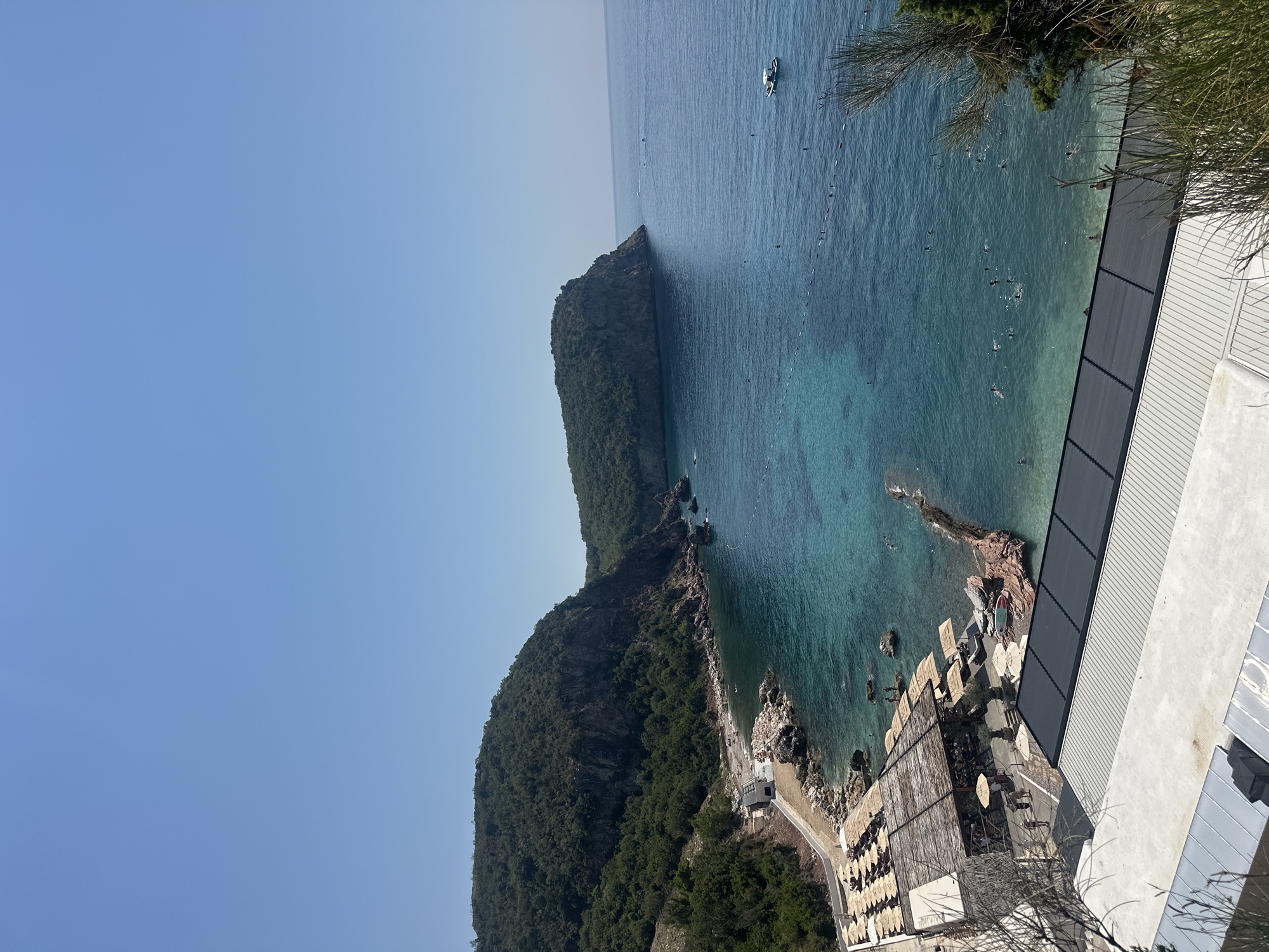 A high-angle, wide shot captures a clear, bright summer day at a coastal resort in Sveti Stefan, Montenegro. The scene unfolds along a rocky shoreline and a turquoise bay. In the foreground, a modern building with a grey, corrugated roof dominates the bottom of the frame, with another section of white, flat roofing above it. Beyond this, a beach area is visible, furnished with rows of evenly spaced wooden sun loungers and umbrellas, suggesting a well-maintained resort. A rustic, open-air structure with a thatched roof serves as a central hub on the beach, with people visible walking around it.
The bay itself is a striking feature, with water transitioning from crystal clear near the shore to a deeper blue further out. Several individuals are swimming and enjoying the water, marked by buoys and ropes delineating a swimming area. A small white boat is anchored in the bay, adding to the sense of leisure.
The background is defined by two imposing, tree-covered hills that rise dramatically from the coastline. The larger hill, on the left, features steep, rocky cliffs meeting the water, while the smaller hill, further out in the bay, forms a prominent headland. The sky is a seamless expanse of clear, light blue, indicating fair weather. The overall impression is one of a serene and picturesque coastal retreat.