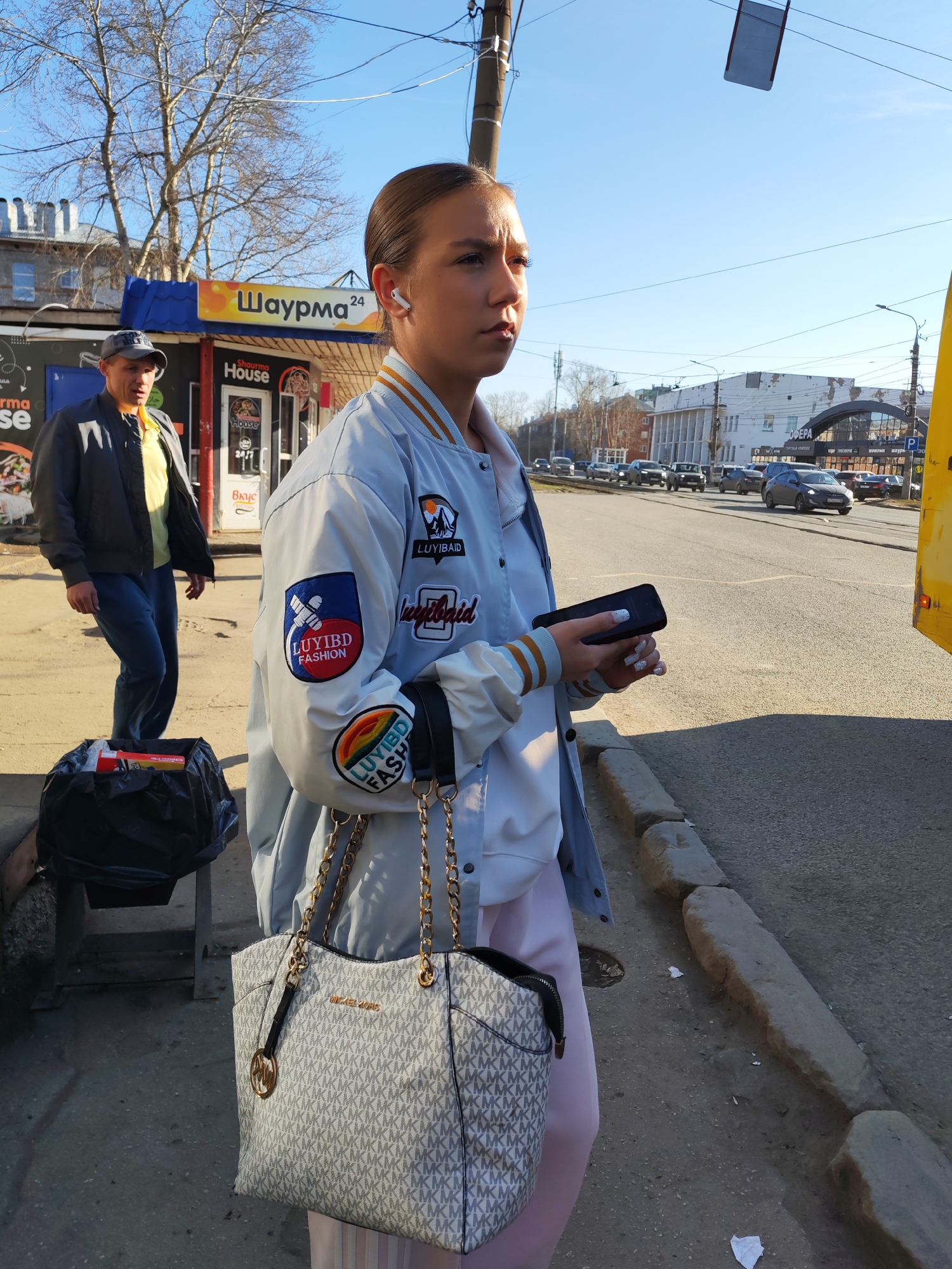 An outdoor, eye-level shot captures a young woman and a street scene in Izhevsk, Russia, under bright, clear daylight. The woman, positioned on the right side of the frame, looks left with a focused expression. She wears a light blue bomber jacket adorned with several patches, including one featuring a mountain scene with "LUYIBD", another with a rocket-like emblem stating "LUYIBD FASHION", and a rainbow patch with "LUYIBD FASH". She has light brown hair pulled back, white wireless earbuds, and holds a black smartphone in her hands. A large white and gray Michael Kors tote bag, with its distinctive monogram pattern and a gold chain strap, hangs from her shoulder. Light pink pants are visible beneath her jacket.
Behind her, to the left, a man in a dark jacket, yellow polo shirt, and baseball cap walks towards the background, carrying a black bag filled with items, possibly packaged goods, near a small stand. The setting is an urban street with bare trees suggesting late autumn or early spring. In the background, across the street, cars are parked and moving. Buildings line the street, including one with "СФЕРА" (SPHERE) visible on its facade and a "P" parking sign.
Prominent signage in the mid-ground includes a bright yellow "Шаурма 24" (Shaurma 24) sign, along with "Shaurma House" and "House," indicating fast-food establishments. A partial yellow structure, likely a tram or bus, is visible on the far right. The overall atmosphere is that of a bustling city street on a sunny day.