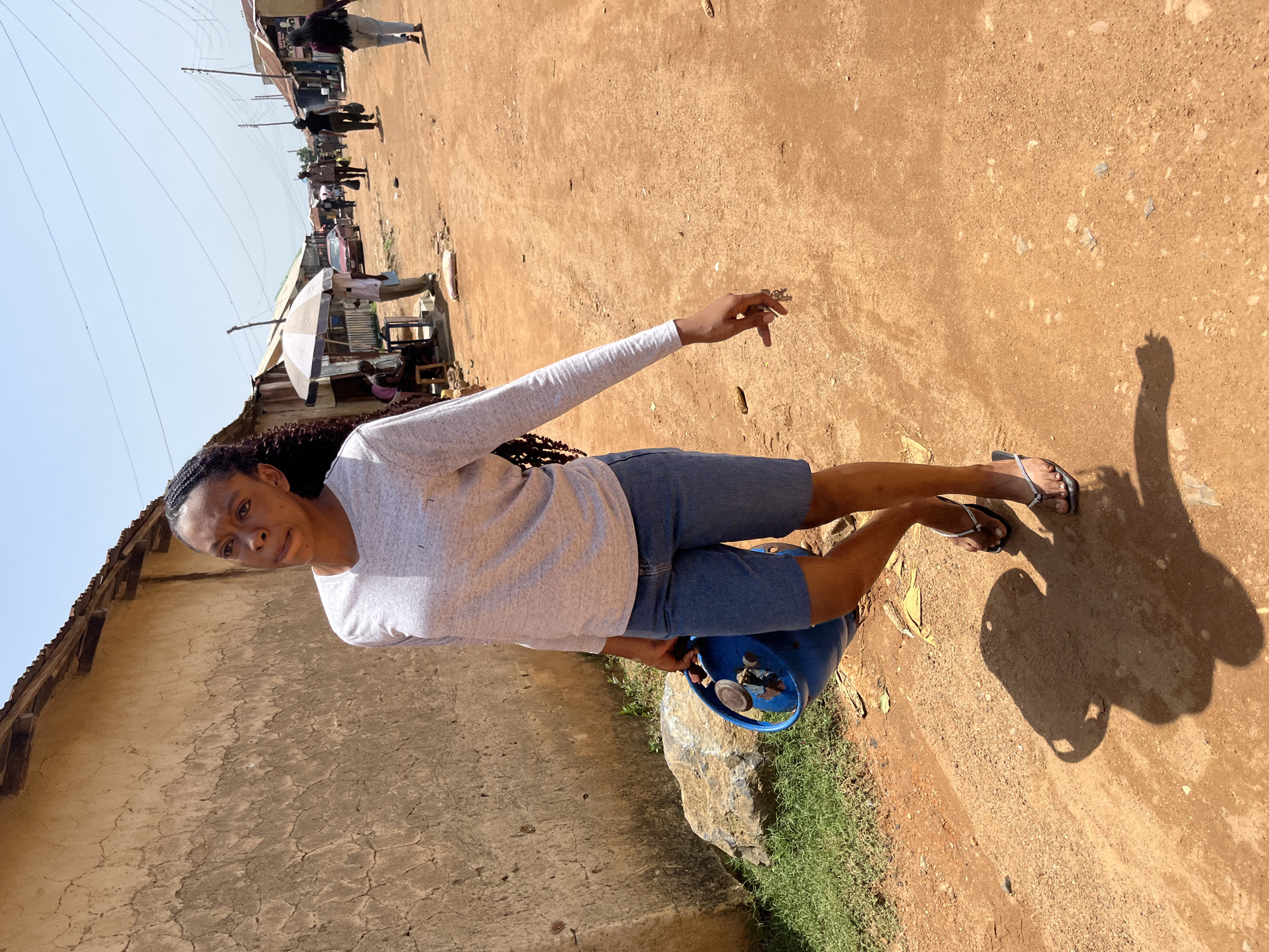 A person, with braided dark hair, wears a light grey long-sleeved shirt and denim shorts. They are standing on a dirt road with a blue gas cylinder at their side. The person's left hand is raised and appears to be holding something small. In the background, there are buildings with earthen walls and corrugated metal roofs, suggesting a rural or semi-urban setting. Several other people are visible in the distance, as well as market stalls with an umbrella. The ground is dusty and a few rocks are scattered around. The lighting suggests it is daytime with the sun casting shadows. The overall atmosphere appears to be lively, as indicated by the presence of multiple people and market activity.