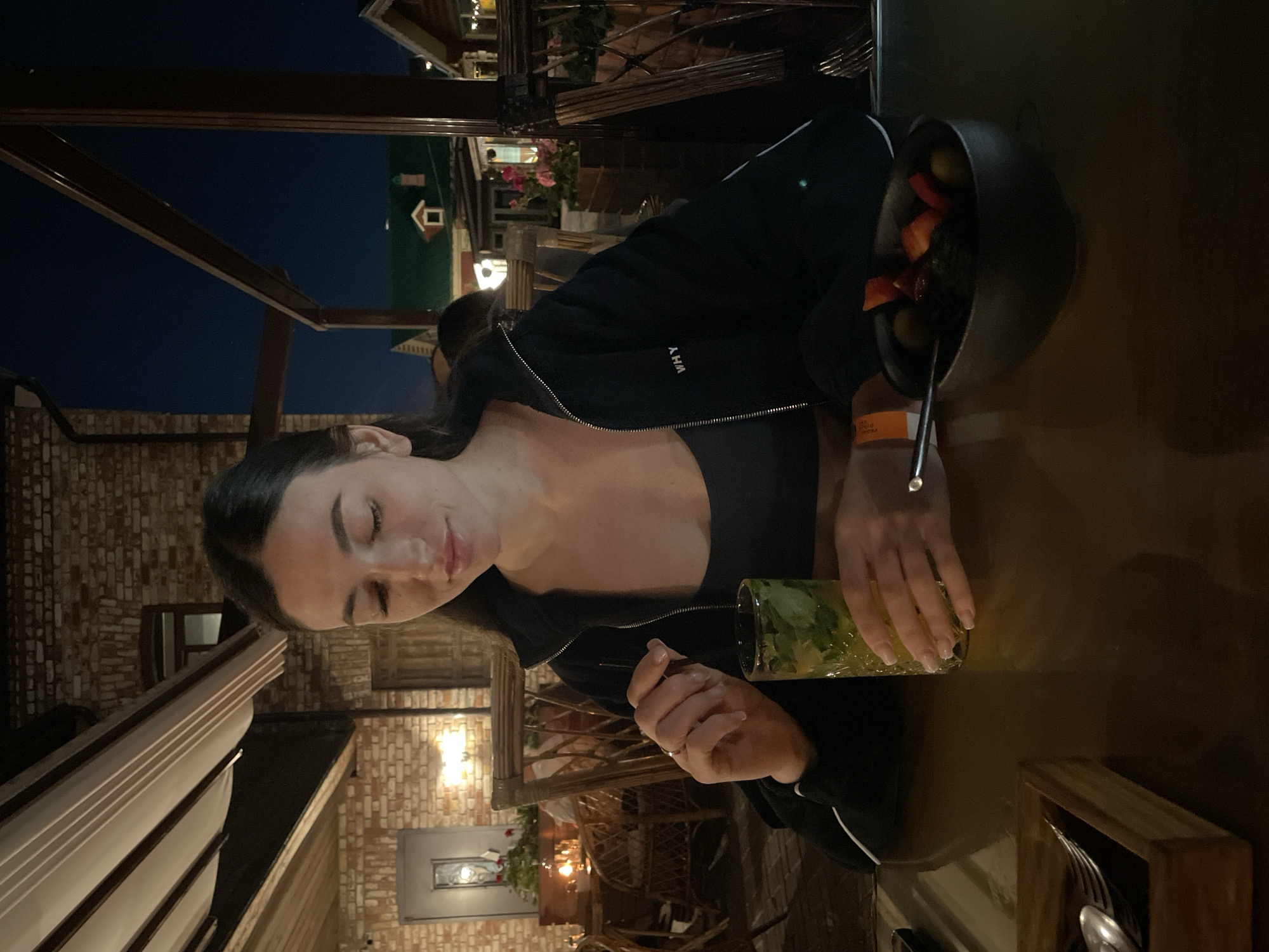 A young woman is sitting at a table in an outdoor restaurant in Kazan, Russia. She wears a black jacket with a white stripe on the sleeve and has long, dark hair. She is holding a glass with green leaves in it, likely a cocktail. A black pot of food is on the table next to her. A wooden box with silverware is also on the table. In the background, there are brick buildings with lights, plants, and other restaurant seating. The sky is dark, indicating it is evening.