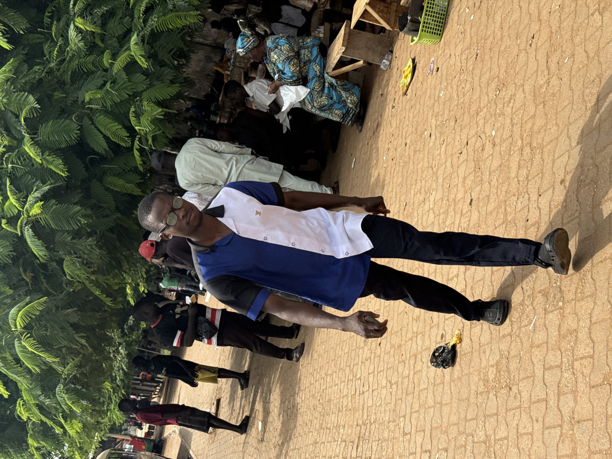 In this image, a young man with glasses is walking towards the camera on a paved surface. He is wearing a blue and white short-sleeved shirt and black pants. Behind him, other people are visible, some standing and others seated. To the left, a red bus is partially visible. The environment appears to be an outdoor marketplace with trees providing shade. The ground is covered with interlocked brick pavers. In the foreground, a black plastic bag is on the ground. In the background, to the right, a woman in a blue and yellow patterned outfit is seated on a wooden bench. There are also some baskets and other items on the ground. The scene is consistent with an urban setting in Abuja, Nigeria, likely a busy area such as a market or transport hub.