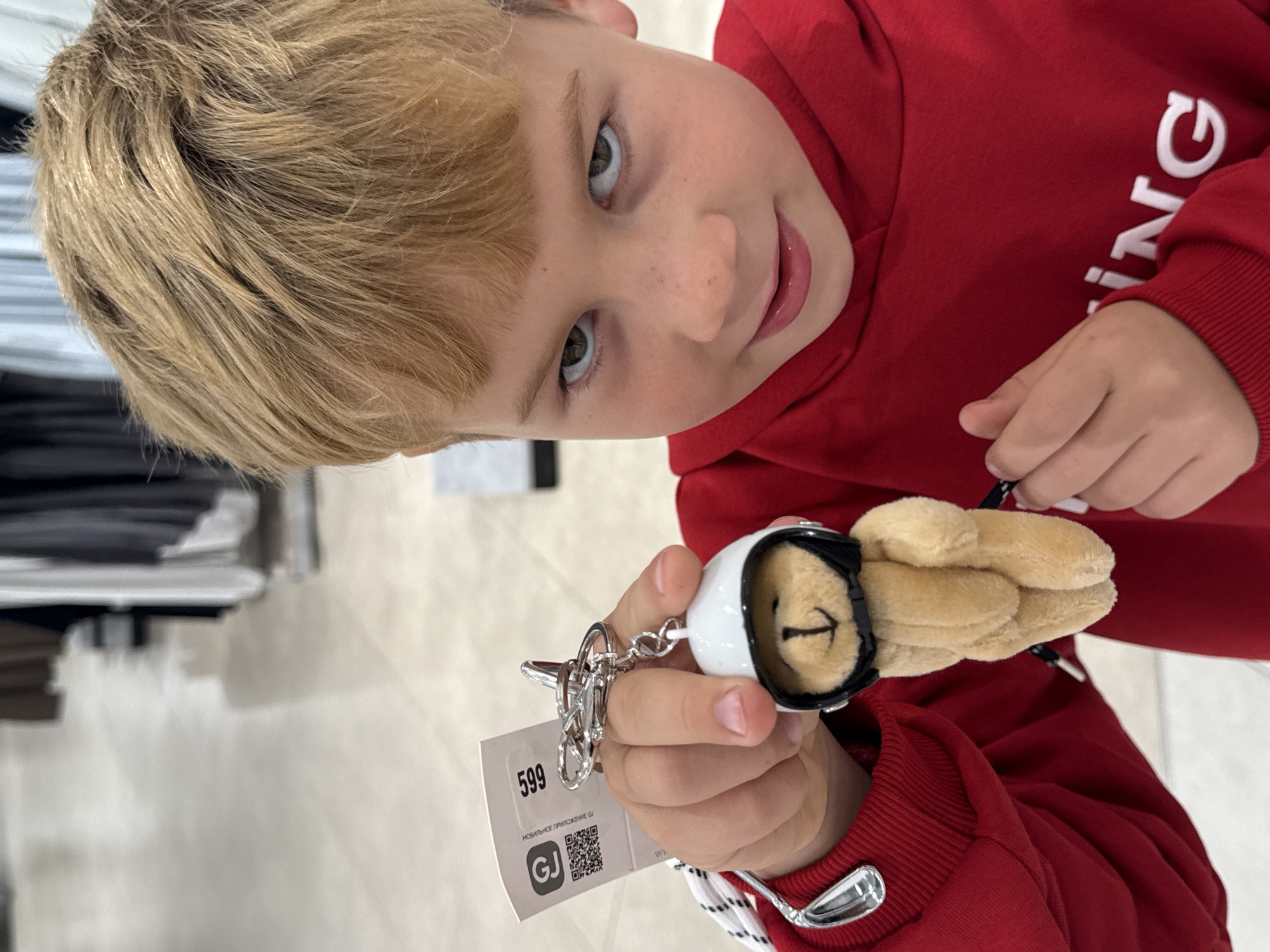A young boy with blond hair and fair skin is holding a teddy bear keychain. The bear is wearing a white helmet with a black visor. The boy is wearing a red sweatshirt. The tag on the keychain has the number "599" and the text "МОБИЛЬНОЕ ПРИЛОЖЕНИЕ GJ" (MOBILE APPLICATION GJ). The QR code is visible on the tag. The letters "ING" are visible on the sleeve of the sweatshirt. The image was taken in Yekaterinburg, Russia, since "GJ" is the shopping mall Golden Grove in Yekaterinburg.