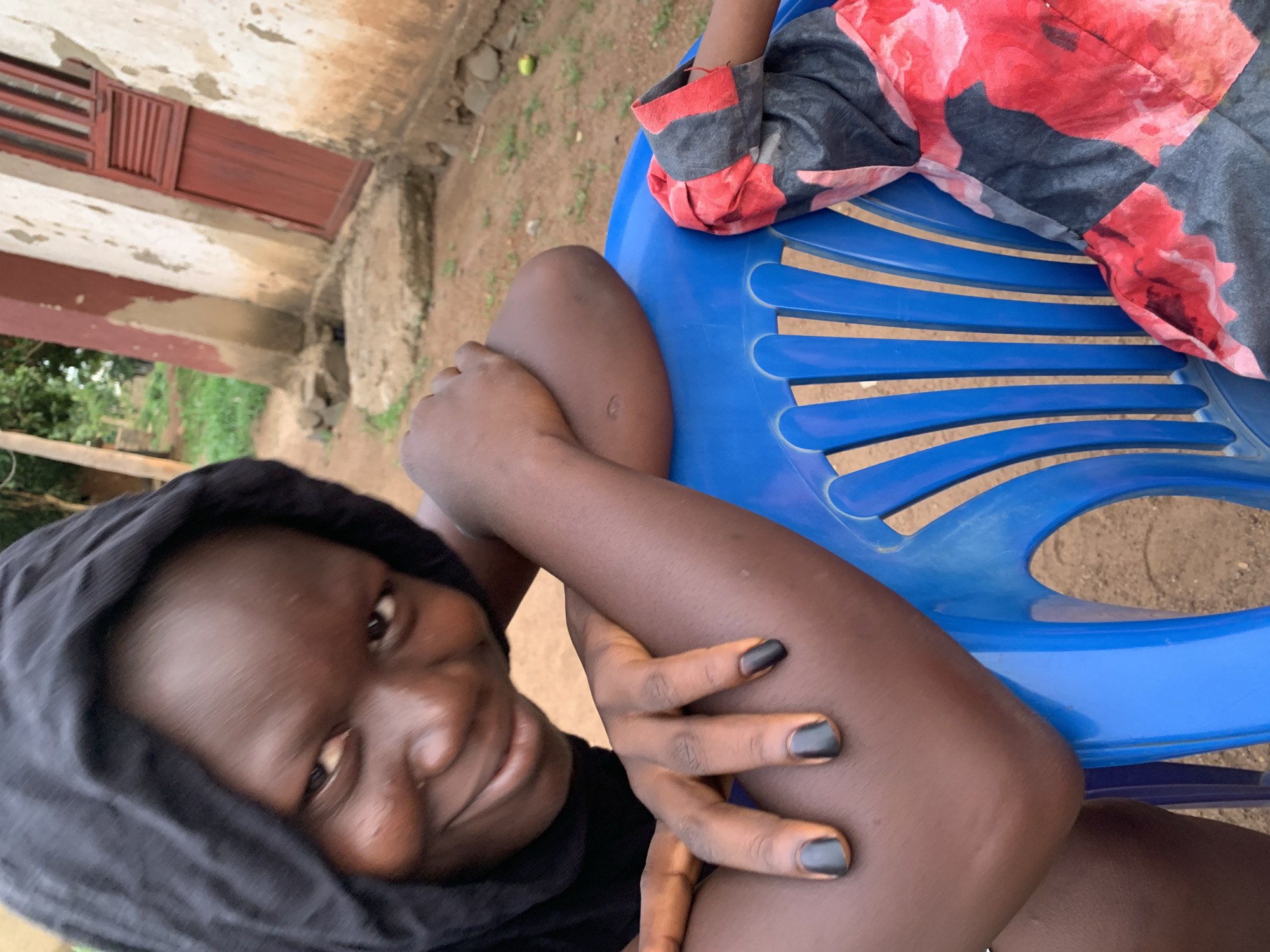 A young Black woman is sitting outdoors in Agban, Nigeria. She wears a black head covering and a black shirt. Her fingernails are painted black. She is sitting on a blue plastic chair, and another person wearing a garment with red and gray floral patterns is partially visible next to her. In the background, a building with a red window frame is visible. The surrounding environment appears to be grassy with dirt.