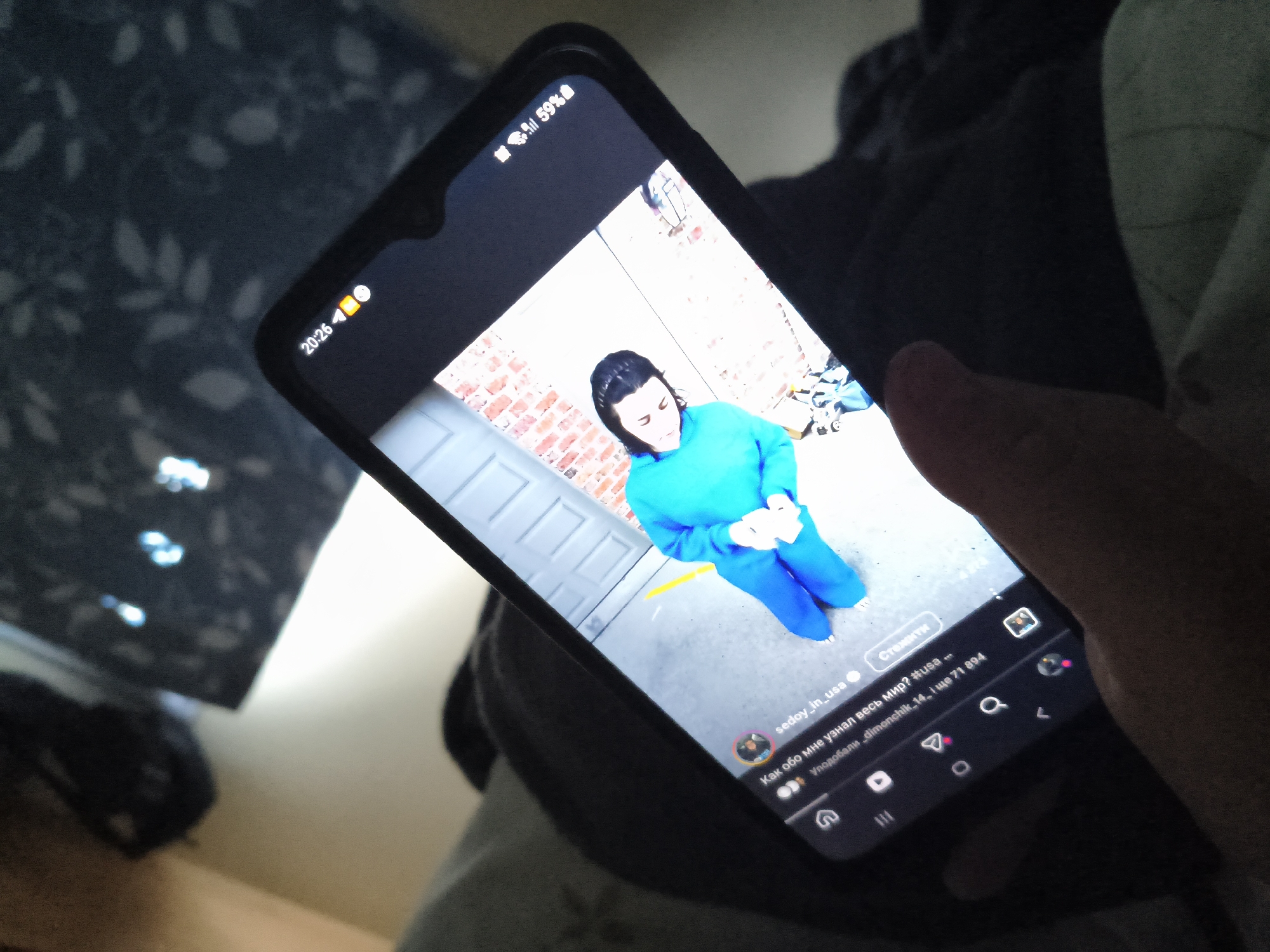 A person's hand is holding a smartphone displaying a social media post. The post features a woman in a bright blue outfit, possibly a tracksuit or jumpsuit. She has dark hair and is looking downwards. The background of the post shows a brick wall and a light-colored garage door. The phone's screen also shows the time "20:26" and a battery indicator showing "59%". The image was taken indoors, with a dark, patterned surface visible in the background, possibly a blanket or upholstery. There is no visible indication of the city of Borowa, Poland.