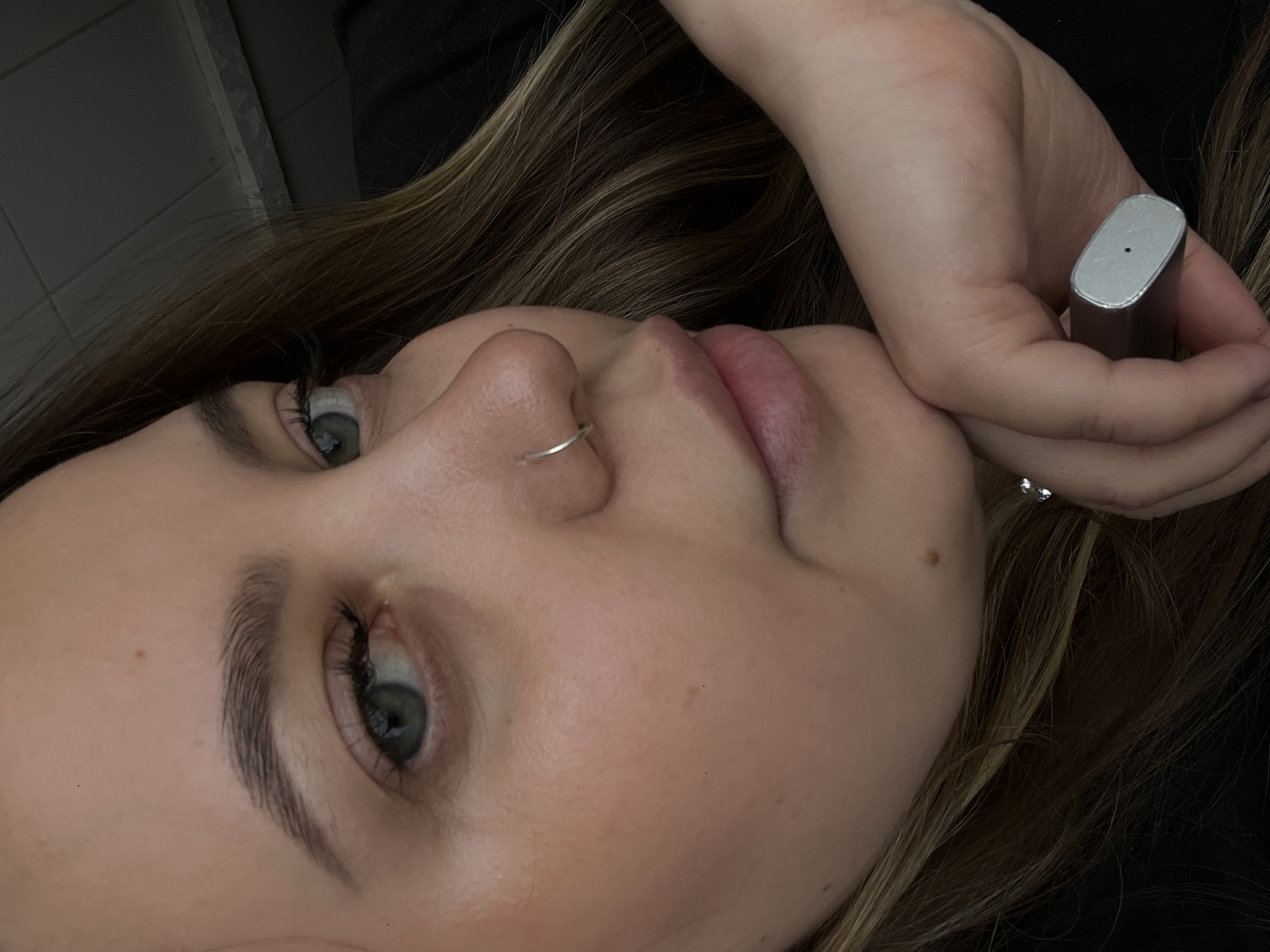 A close-up, angled portrait features a young woman with long, light brown hair, likely in her late teens or early twenties. Her face is positioned prominently, showcasing her fair complexion, light blue-green eyes, and full lips. A small, thin silver nose ring adorns her right nostril. Her gaze is directed subtly to the left side of the frame, conveying a calm, possibly pensive or thoughtful expression.
Her right hand is brought up towards her face, resting gently near her cheek or chin. She holds a small, sleek, silver-grey rectangular object, which resembles a compact electronic device such as a vape pen or a portable charger. A silver ring is visible on one of her fingers.
The background is dark and largely out of focus, suggesting an indoor environment. A segment of a grey tiled wall or surface is faintly discernible in the upper left corner. The lighting softly illuminates her features, drawing attention to her eyes and the details of her face and hand. No visible text is present in the image. This scene is located in Novopokrovskaya, Russia.