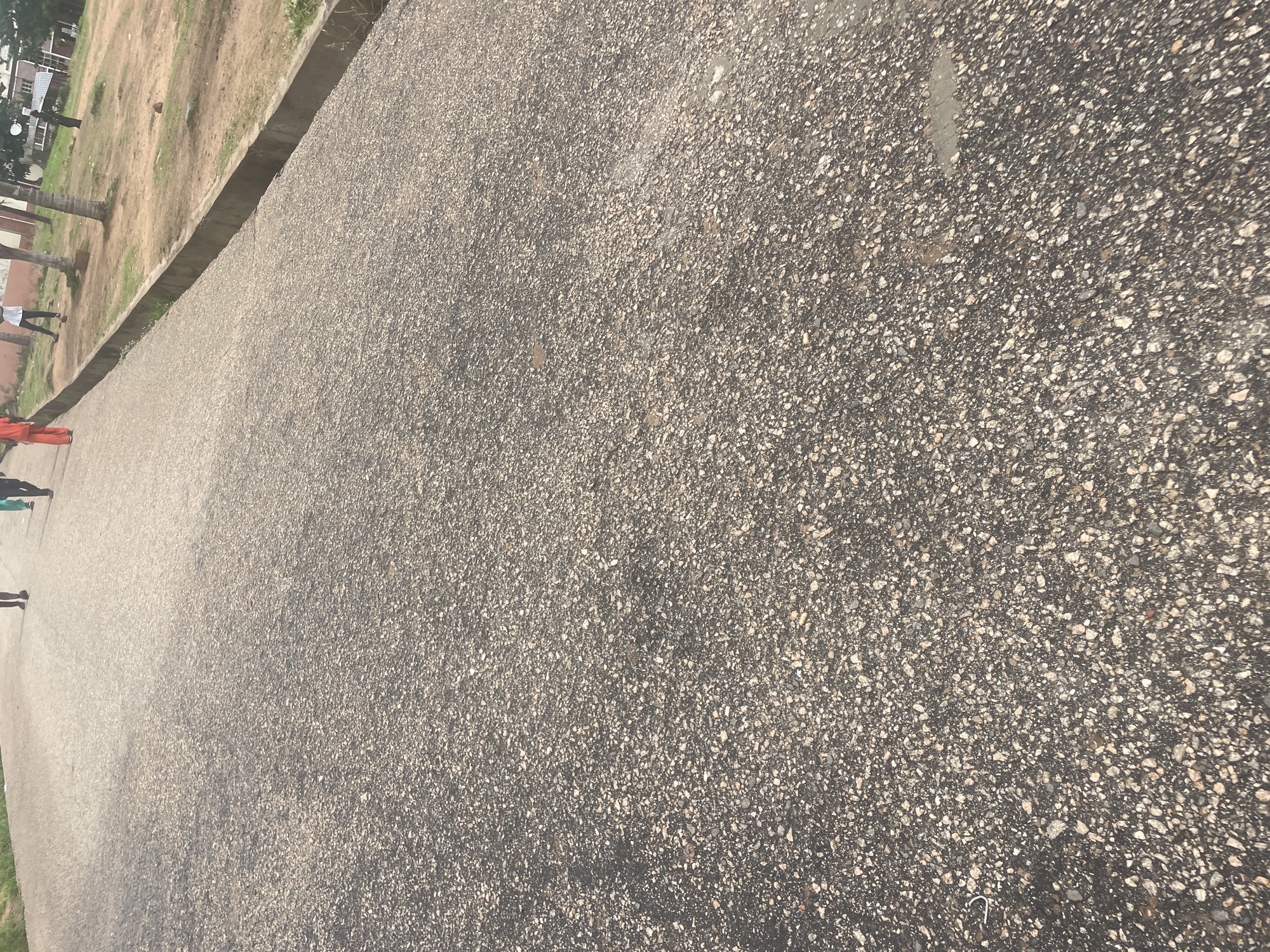 The media file displays an outdoor scene with a focus on a paved surface. The paved area is comprised of small, dark grey pebbles mixed with lighter grey and white stones, giving it a textured, gravelly appearance. A distinct concrete border runs along the left edge of the paved area, separating it from a strip of dry, light brown earth and sparse green grass.
In the background, beyond the earthy strip, a few palm trees are visible, along with hints of buildings with white walls and brown roofs, suggesting an urban or semi-urban environment. On the far left of the image, several people are walking along the paved path. One person is wearing an orange outfit, and another is in dark clothing. The lighting suggests it is daytime, with no strong shadows indicating a specific time of day, and the weather appears dry. The overall impression is a simple, everyday scene in Lokoja, Nigeria. No visible text is present in the media file.