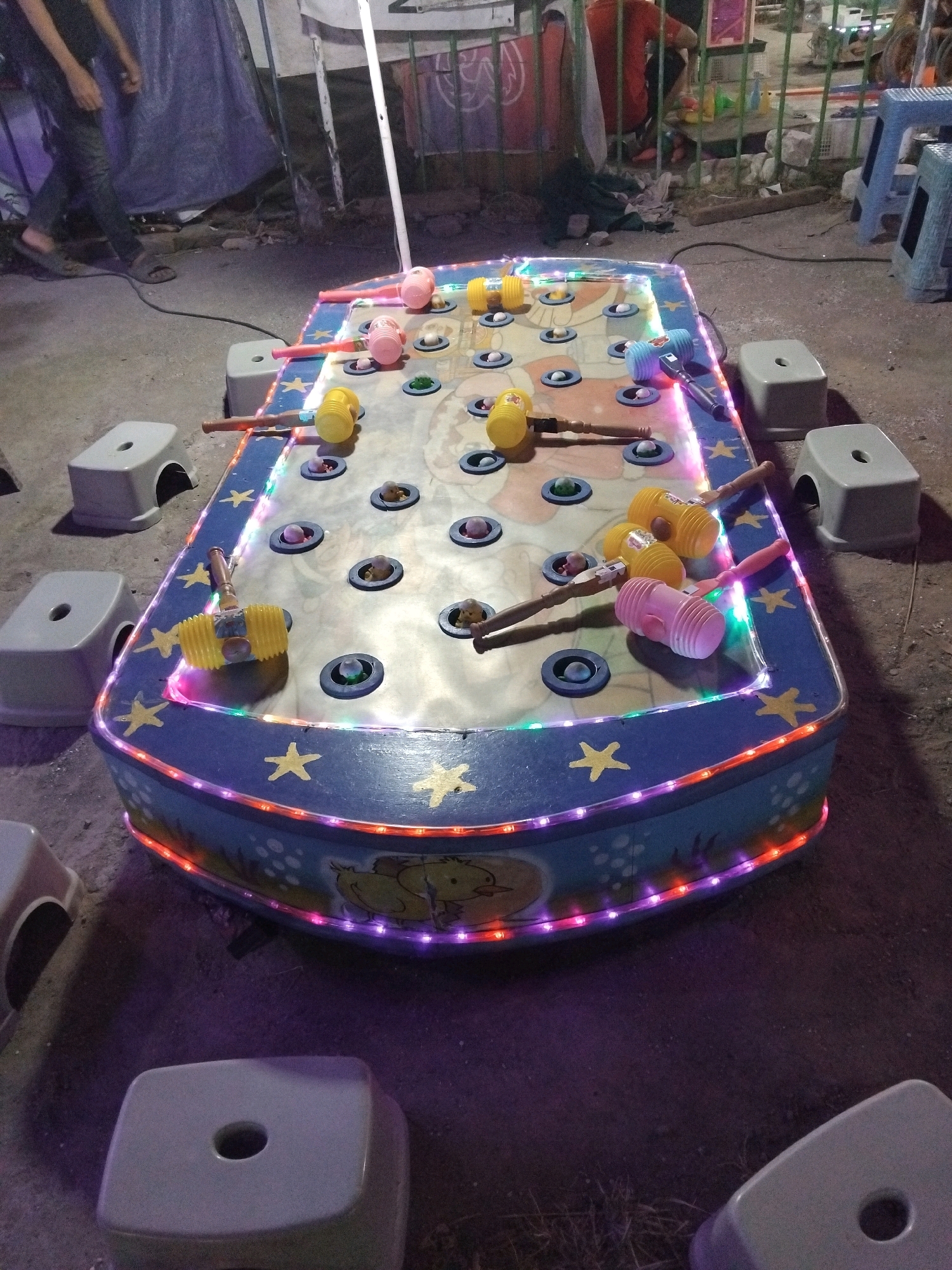 A whac-a-mole style game is set up outdoors in Sedati Agung, Indonesia. The game board is rectangular with a blue and yellow checkered pattern and features several holes where mole-like figures pop up. Several toy hammers, some yellow and some pink, are scattered across the game board. The edges of the game board are decorated with colorful LED lights and yellow stars. Surrounding the game are several small, light gray plastic stools. In the background, a person is walking away from the camera, and a blue tarp is visible. The ground appears to be a mix of concrete and dirt.