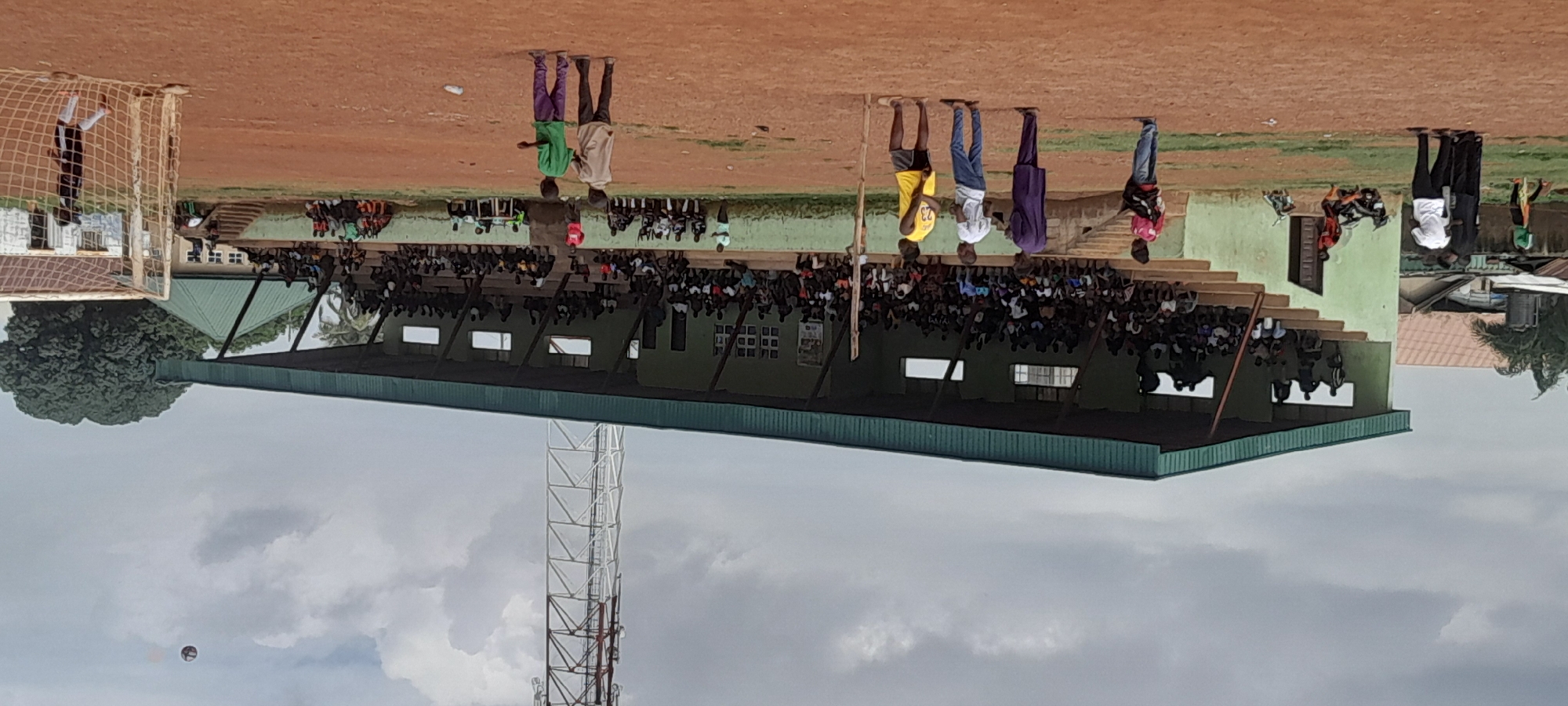 The image shows a football stadium in Abuja, Nigeria, under a cloudy sky. Spectators are seated in a tiered, covered grandstand which is painted light green and has a dark green roof. A tall, white lattice telecommunications tower stands behind the grandstand. On the field, which is a dirt pitch, several people are present. Some are standing, while others appear to be involved in a game. A football is visible in the air in the upper right portion of the image, and a goalpost with a net is also in the right foreground. Buildings with corrugated iron roofs and other structures are visible in the background.