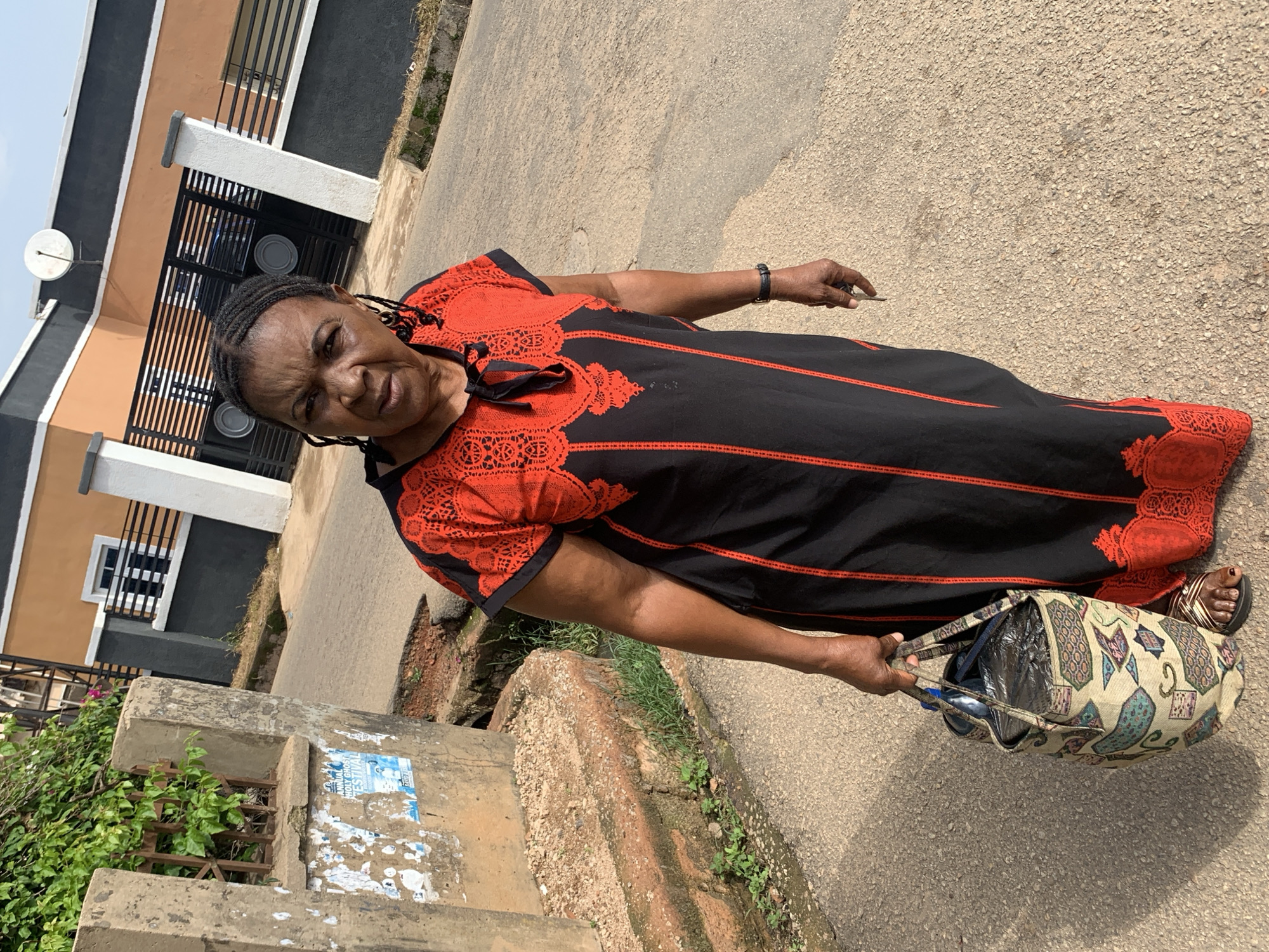 An elderly Black woman with dark skin and braided hair is standing on a paved road in front of a two-story building with a distinct orange and gray facade. She is wearing a black and red patterned dress and carrying a patterned tote bag in her left hand. The building has a dark metal gate with horizontal bars and a white satellite dish mounted on the upper right corner of its orange section. To the left of the woman, there is a concrete structure with posters plastered on it, and overgrown green foliage spilling over a metal grate. The paved road is cracked and has some reddish-brown soil visible. The scene appears to be in an urban or suburban area, likely within Jos, Nigeria, based on the architectural style and vegetation.