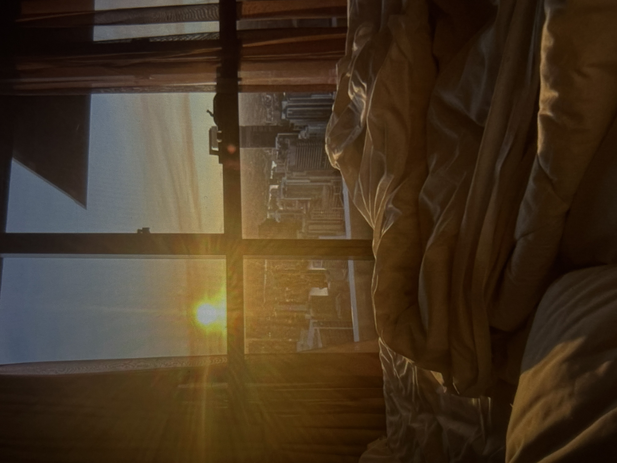A sunlit bedroom with a city view at dawn features an unmade bed in the foreground, with rumpled white and beige sheets and blankets. A large window dominates the background, revealing a panoramic cityscape bathed in the warm, golden light of sunrise. Tall buildings, including modern skyscrapers and older structures, are visible in the distance. Sheer orange curtains frame the window on the right and left sides. The sun is positioned low in the sky, casting strong rays and lens flare across the image. The scene is set in an urban environment, likely from an elevated vantage point within a building. The image does not provide any specific information or context to suggest the location is Knezha, Bulgaria.