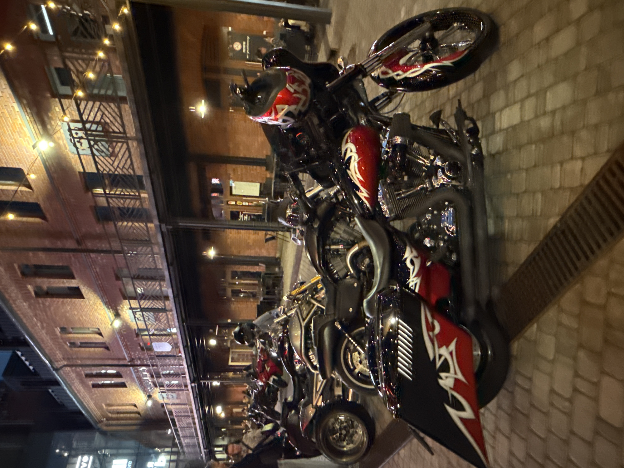 The image depicts a street scene at night, likely in a historic district of Saint Petersburg, Russia, with an emphasis on parked motorcycles. In the foreground, a custom motorcycle with red and white flame decals and a black fairing is prominently displayed. To its left, another motorcycle with a similarly styled red and white decal on its side bag is partially visible. Behind these, more motorcycles are parked, including a red one and a darker one with a helmet resting on its handlebars.
The background features a multi-story brick building illuminated by string lights and outdoor lamps, suggesting a commercial or entertainment area. Balconies with decorative railings line the upper floors. Several figures are visible, mostly out of focus, some appearing to be patrons or staff of establishments along the street. The ground is paved with cobblestones and features a metal grate drain. The overall ambiance is one of an evening gathering or event centered around the motorcycles.