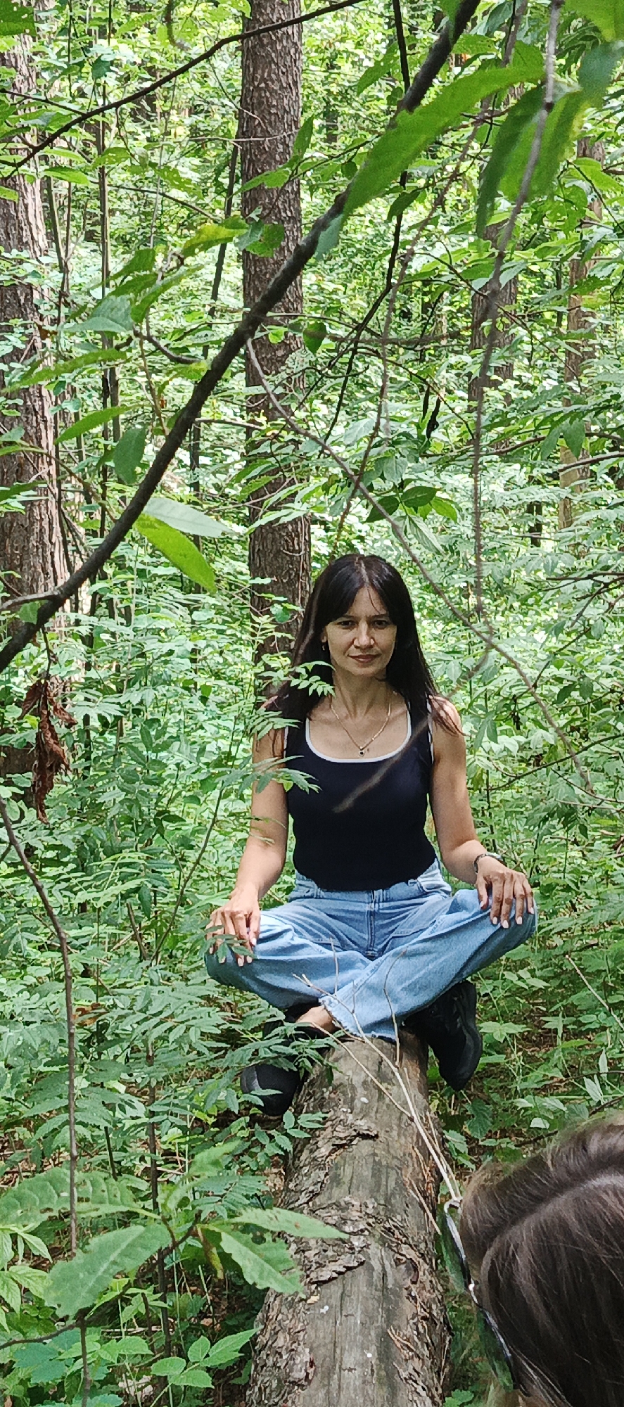 A woman with long, dark hair is meditating in a forest, seated cross-legged on a fallen log. She is wearing a black tank top with white trim and light-wash, wide-leg jeans. A delicate necklace with a dark pendant adorns her neck. Her hands are resting on her knees, with her fingers loosely curled. The forest is lush and green, with trees and dense foliage surrounding her. Sunlight filters through the leaves, suggesting it is daytime. The overall mood is serene and peaceful. The image also shows a partial view of another person's head and shoulders in the foreground, to the right of the frame, with dark hair and glasses. The location is identified as Chaikovsky, Russia. There is no visible text in the image.