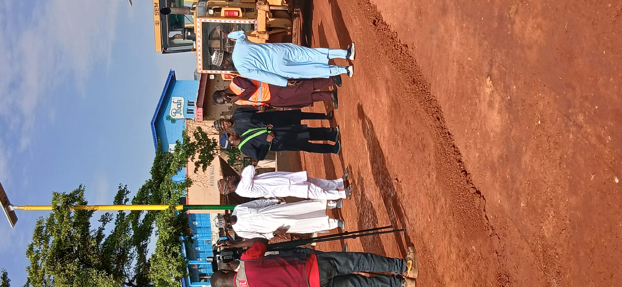 The image depicts an outdoor scene in Zaria, Nigeria, likely in an area undergoing construction or renovation, as indicated by the red earth ground and visible tire tracks. In the background, a blue building with "Peak" branding and palm tree imagery is visible, along with a yellow SEM heavy machinery vehicle.
In the foreground, a group of men are gathered, appearing to be in conversation or observation. One man in a red shirt is filming with a professional camera on a tripod. Several other men are dressed in traditional Nigerian attire, including white and blue kaftans and caps. One individual wears a bright orange high-visibility vest over a maroon garment, suggesting a role in supervision or safety. Another man is wearing a dark cap and a reflective green vest, possibly indicating a security or traffic management role. The overall atmosphere seems to be one of inspection or a site visit. The sky is clear and blue with some clouds.