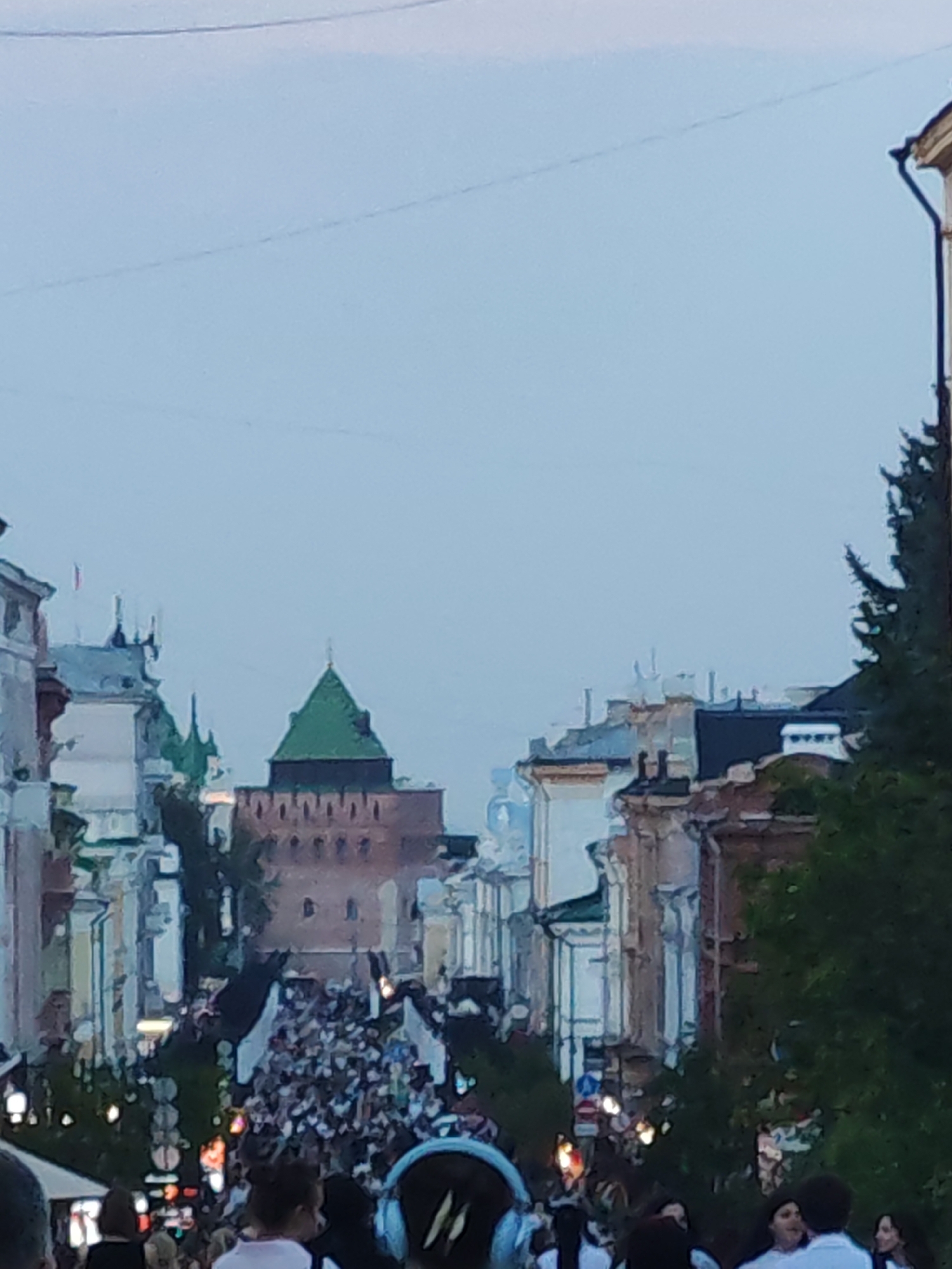 The image captures a street scene in Nizhny Novgorod, Russia, likely during an event or gathering. In the background, a distinct red brick tower with a green pyramidal roof stands prominently, characteristic of the Nizhny Novgorod Kremlin. The tower is surrounded by other historical buildings with varying architectural styles, some featuring arched windows. A dense crowd of people is visible in the middle ground, filling the street and appearing to be moving in a procession or gathered for an event. Many individuals are holding white flags or banners. The foreground shows glimpses of people, some wearing headphones, and the tops of buildings and trees. The overall atmosphere appears to be that of a public event in an urban historical setting under a pale, overcast sky.