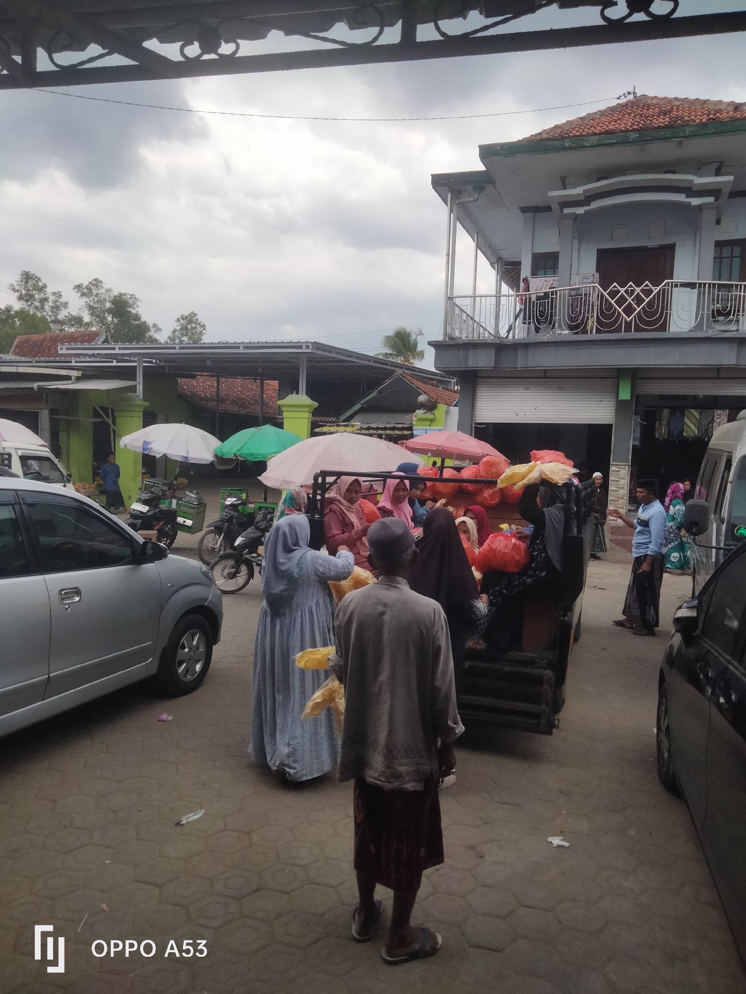 The image captures an outdoor scene, likely a market or a loading area, under an overcast sky. In the foreground, a silver car is parked on a paved ground with hexagonal tiles. To the right, a portion of a black car is visible. A man in a light gray shirt and patterned sarong stands facing away from the camera, holding a yellow plastic bag.
Behind the man, a flatbed truck is partially filled with people and numerous bright orange plastic bags, presumably merchandise. Several individuals, mostly women wearing hijabs and traditional clothing, are interacting around the truck. Some are reaching into the truck, while others are talking. Three umbrellas, one white, one green, and one pink, are set up near the truck, providing shade.
In the background, there are several buildings. To the left, a low-rise green building with a corrugated metal roof is visible, with a person in a blue shirt standing outside. Further back, trees can be seen. To the right of the truck, a two-story building with a red tile roof and a white balcony is prominent. The overall atmosphere suggests a busy, casual environment, possibly a local distribution point for goods. There is no specific visual information in the image that allows for precise identification of the location as Badung, Indonesia.
