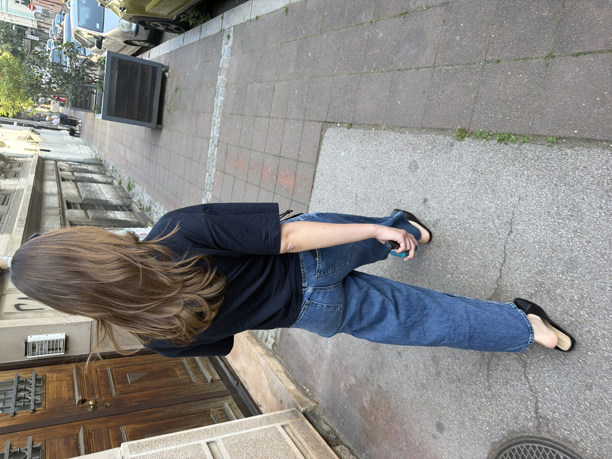 A woman with long brown hair, wearing a black t-shirt, blue jeans, and black flats, is walking on a sidewalk. She is holding a small, rectangular object in her right hand. The sidewalk is made of gray concrete and is bordered by a street with parked cars on one side and buildings on the other. The buildings are made of stone and have wooden doors and windows. The scene is likely in Belgrade, Serbia, given the architectural style.