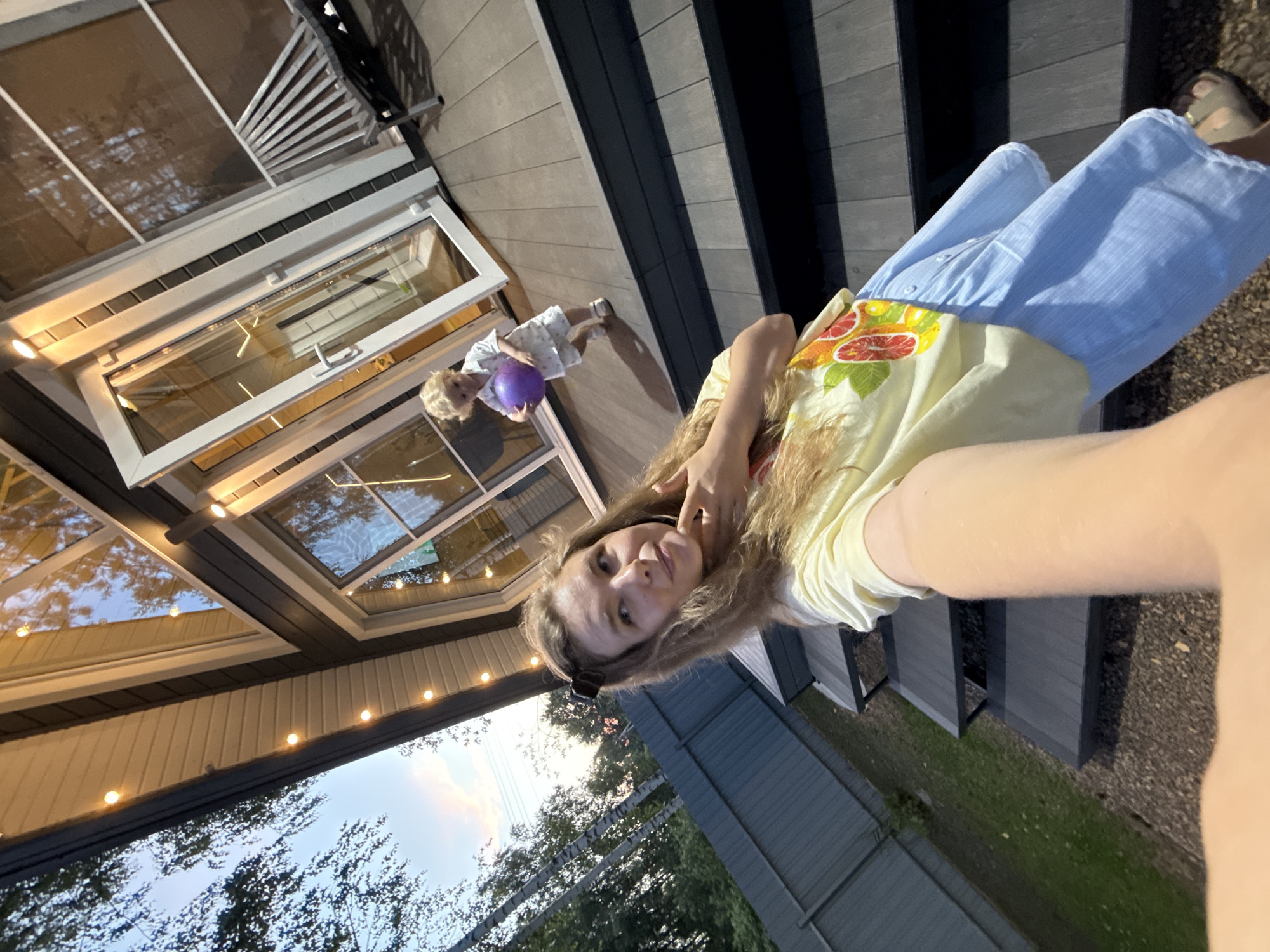 This is a vertical, slightly angled, outdoor selfie taken by a young woman in Prokudino, Russia. She is wearing a light yellow t-shirt with a colorful fruit print and a light blue skirt. Her long, blonde hair is loose, and she is looking at the camera with a relaxed expression, her hand resting near her face.
In the background, a young child with blonde hair is visible, wearing light-colored clothing and holding a purple ball. The child is standing near a large glass sliding door, which appears to be part of a house or building with a modern design, featuring a glass roof. The exterior of the building has wooden-like siding.
The scene appears to be a backyard or patio area, with wooden decking and some grey fencing visible. String lights are hung along the edge of the building, suggesting it might be evening or dusk, and the sky in the background shows a twilight glow. The overall atmosphere seems casual and familial.