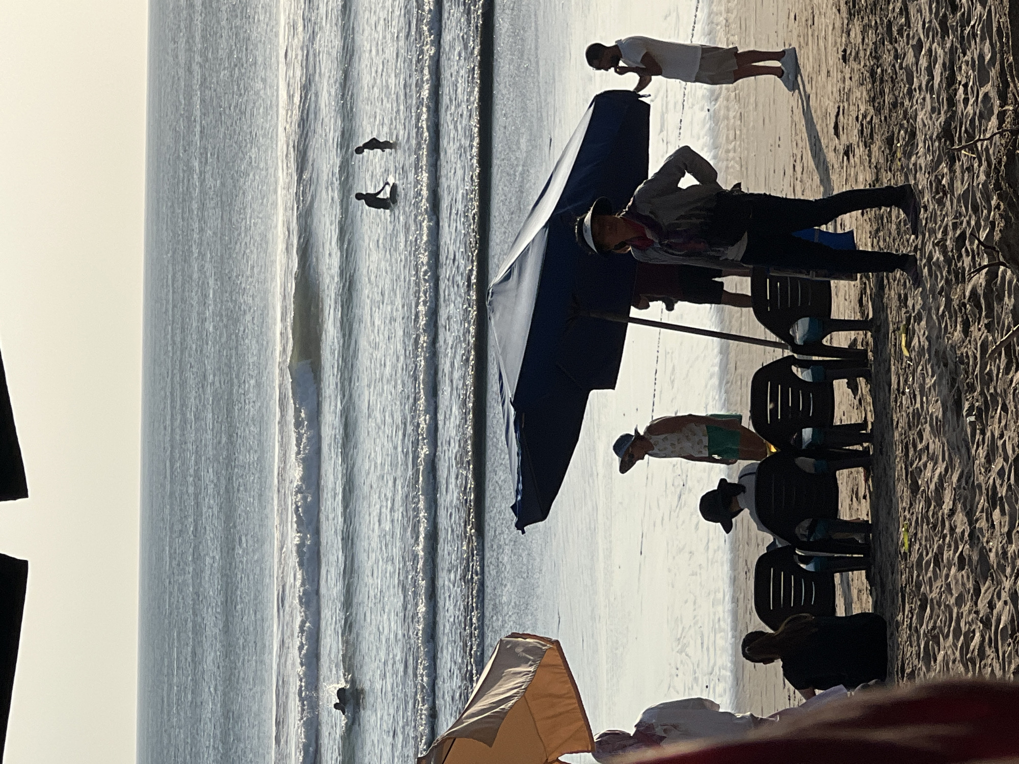 The image depicts a beach scene in Kuta, Indonesia, during what appears to be late afternoon, given the lighting and long shadows. In the foreground, on the sandy beach, several people are visible. Two individuals are seated in dark plastic chairs, facing away from the camera, with one person appearing to have dark hair and the other wearing a dark hat. To the right of them, a woman in a hat, scarf, and dark pants stands looking towards the left. Further to the right, a man in a white t-shirt and shorts stands, looking out at the water. Two large beach umbrellas, one orange and one dark blue, are positioned on the sand, providing shade. In the background, the ocean features moderate waves, and several people are in the water. Two figures are in the shallow water near the shore, one appearing to be on a surfboard. Another person is further out in the water, seemingly swimming. The horizon is visible with a clear sky above the ocean.