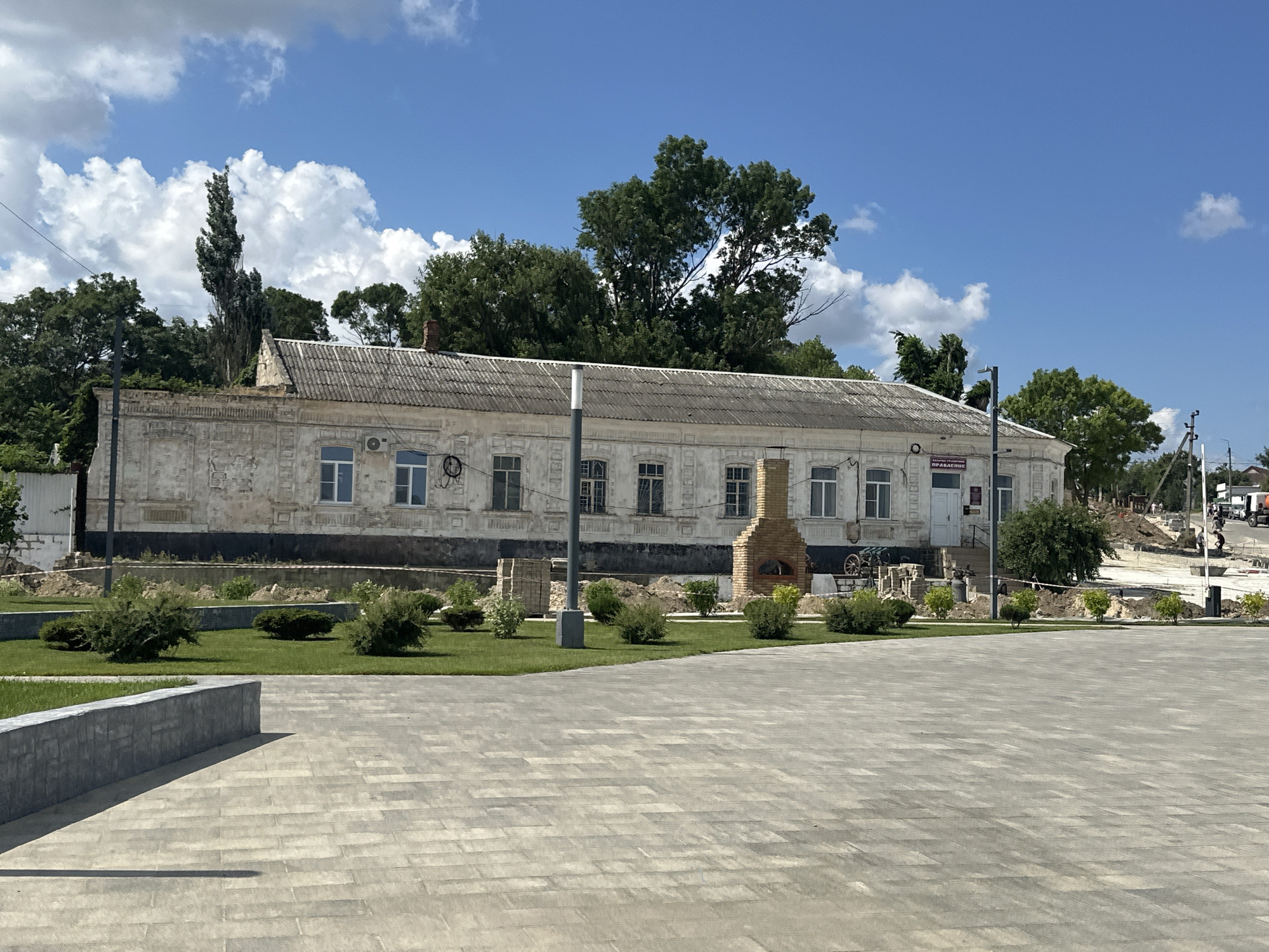 The image depicts a long, low-lying, historical building with a worn, textured facade, located in the city of Taman, Russia. The building has a grey, corrugated roof and a series of regularly spaced windows with white frames. In front of the building, there is a paved plaza with a patterned surface. A low, grey stone wall runs along the edge of the plaza, and beyond that, a well-maintained lawn with several small, rounded bushes.
To the right of the building, a prominent brick chimney with a rounded opening at its base, possibly a pizza oven or a forge, is visible. Several pieces of construction material, including stacks of bricks and wooden beams, are placed in front of the building.
In the background, lush green trees form a dense canopy above the building. Beyond the trees, a bright blue sky with scattered white cumulus clouds dominates the upper portion of the image. A tall, slender tree with a dark, textured trunk stands out against the sky to the left of the building.
Further in the background, to the right, the scene appears to be a construction or renovation area with disturbed earth and visible infrastructure, including what look like utility poles and a truck. The overall impression is a blend of historical architecture and ongoing development.