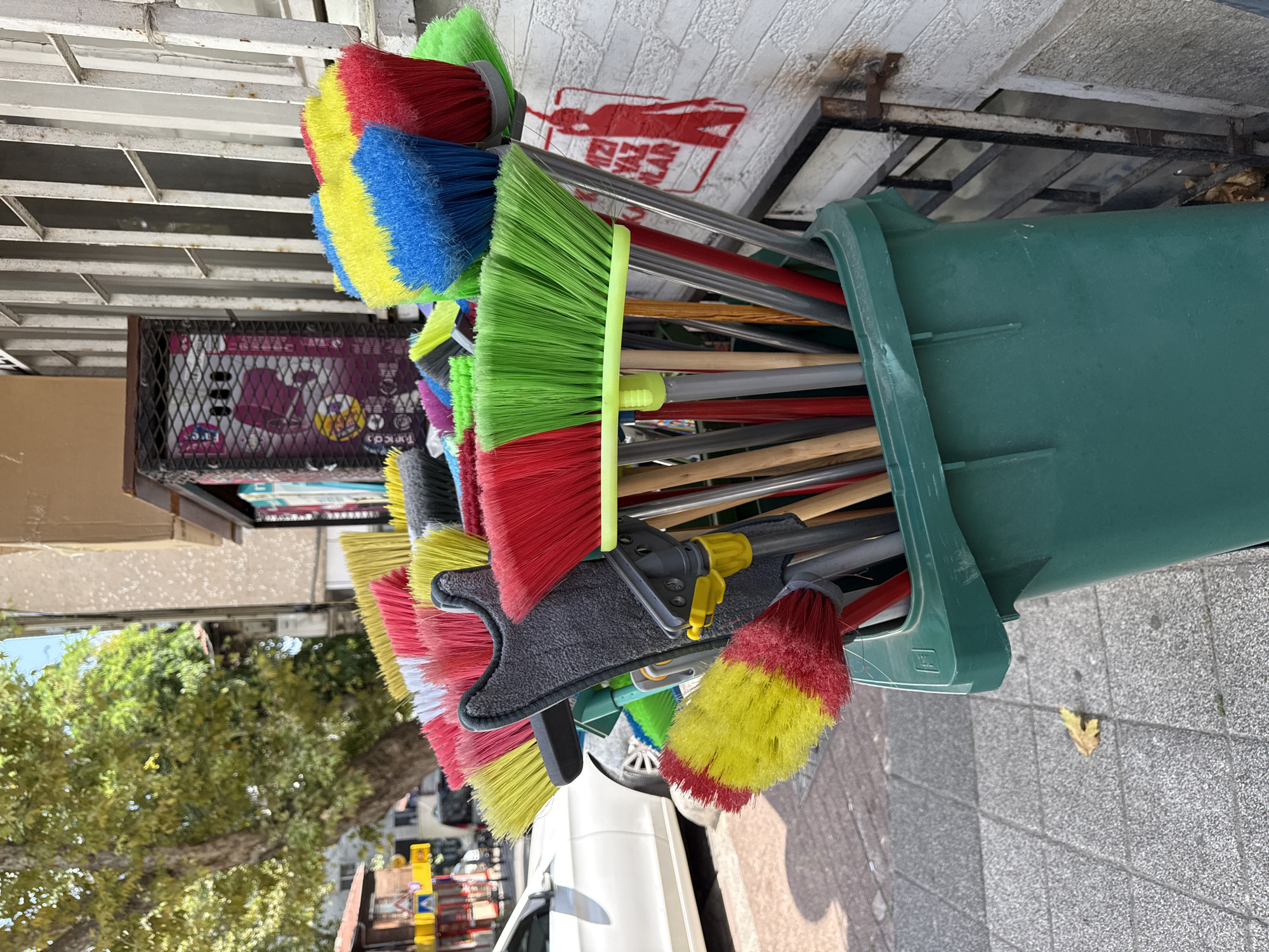 In Kadıköy, Turkey, a collection of brightly colored brooms and cleaning tools are displayed in a large green plastic bin on a sidewalk. The brooms have bristles in red, yellow, blue, and green, with some also having grey or white. The bin is positioned near a white brick wall with a metal gate and a red silhouette graphic of a person. Behind the bin, a white car is parked on the street, and a large tree with green foliage is visible. The ground is paved with grey tiles, and a fallen yellow leaf is present on the pavement. A metal grate with a box behind it is also in the background. The scene appears to be an outdoor display of cleaning supplies, likely outside a shop.