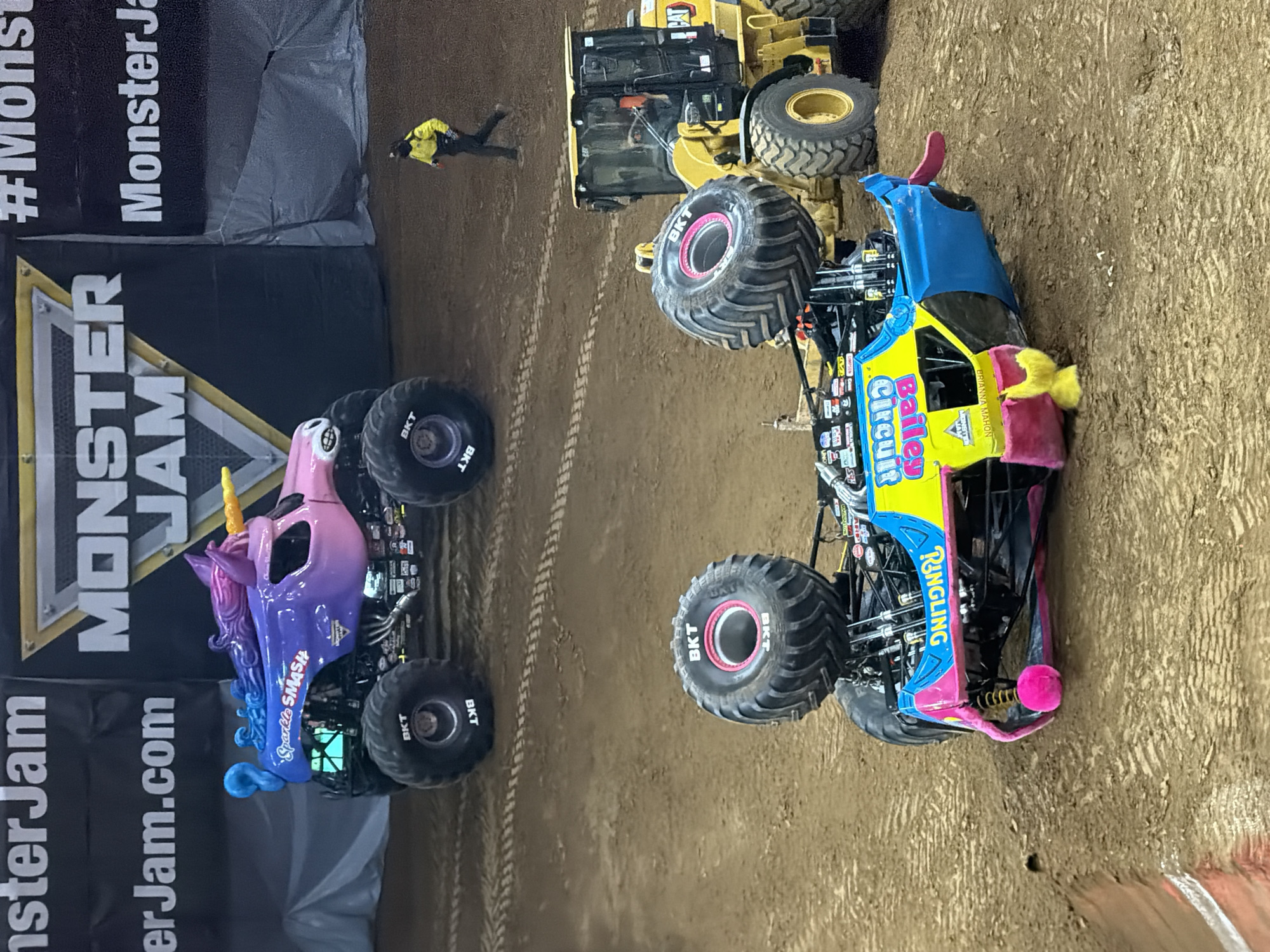 In Sacramento, California, on dirt terrain, two monster trucks are present. One truck, "Sparkle Smash," is upright and decorated with a unicorn theme, featuring purple, pink, and blue colors. It has large black tires with "BKT" branding. The second truck, "Bailey Circuit," is overturned on its side and displays bright yellow, blue, and pink colors, with "BAILEY CIRCUIT" written on its side in yellow text with a pink outline. A bright pink pom-pom is attached to the front underside of this overturned truck. A yellow loader vehicle is parked behind the overturned "Bailey Circuit" truck. A person wearing a yellow vest and black pants is walking away from the two monster trucks towards the background, where a large banner with "MONSTER JAM" logo is visible. The environment appears to be an indoor arena or stadium, indicated by the presence of the banner and lighting, though the dirt ground suggests an event taking place on a prepared surface.
